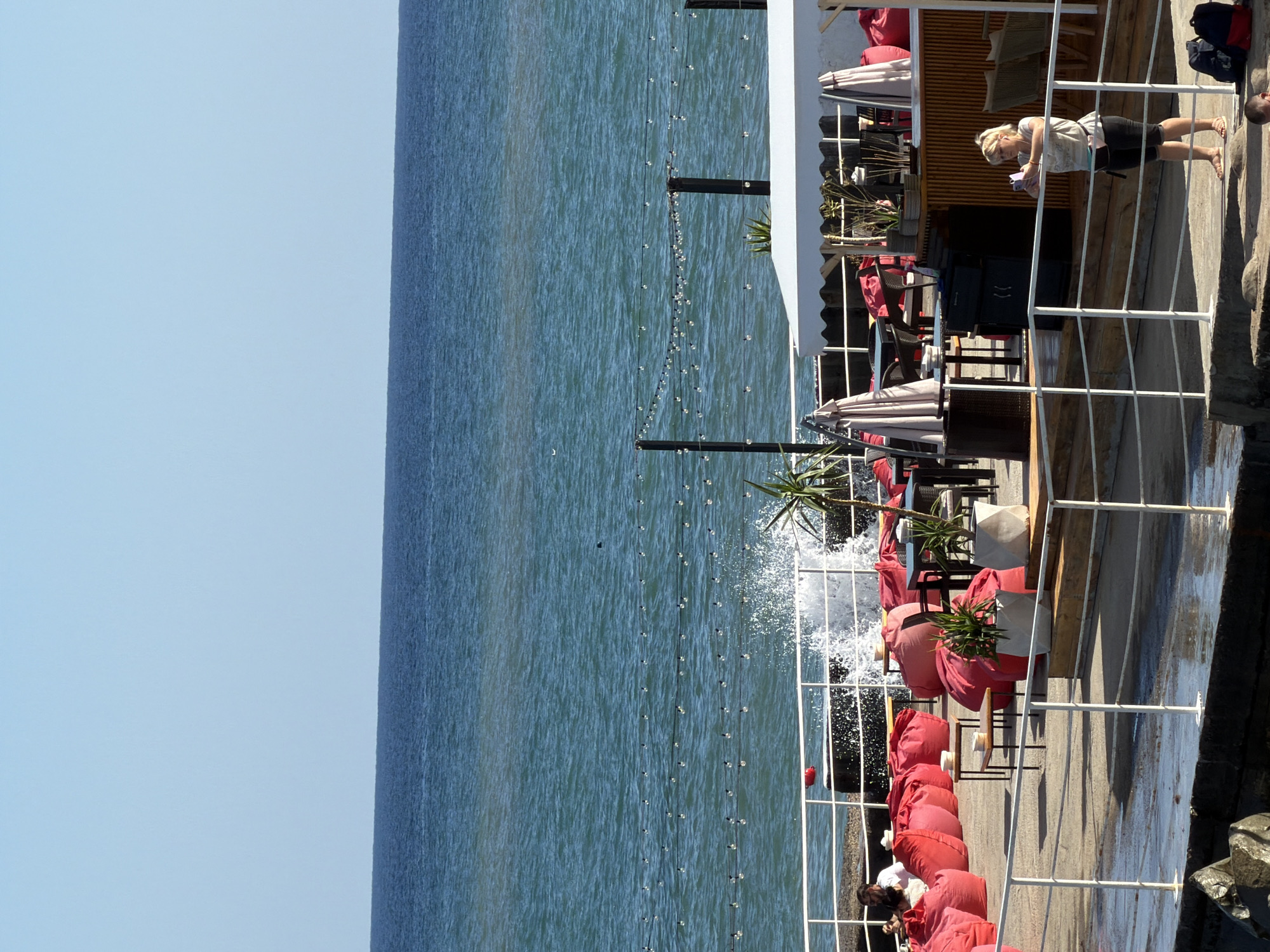 The image depicts an outdoor seating area near the sea, likely a cafe or restaurant, with a person in the foreground and a woman standing by a railing, looking at her phone. The scene is set against the backdrop of the blue sea and sky. In the midground, there are numerous red beanbag chairs, tables, and umbrellas, suggesting a relaxed atmosphere. A wave crashes against a wall, creating a spray of water. In the background, the sea stretches to the horizon under a clear blue sky. The overall impression is of a sunny day at a seaside establishment.