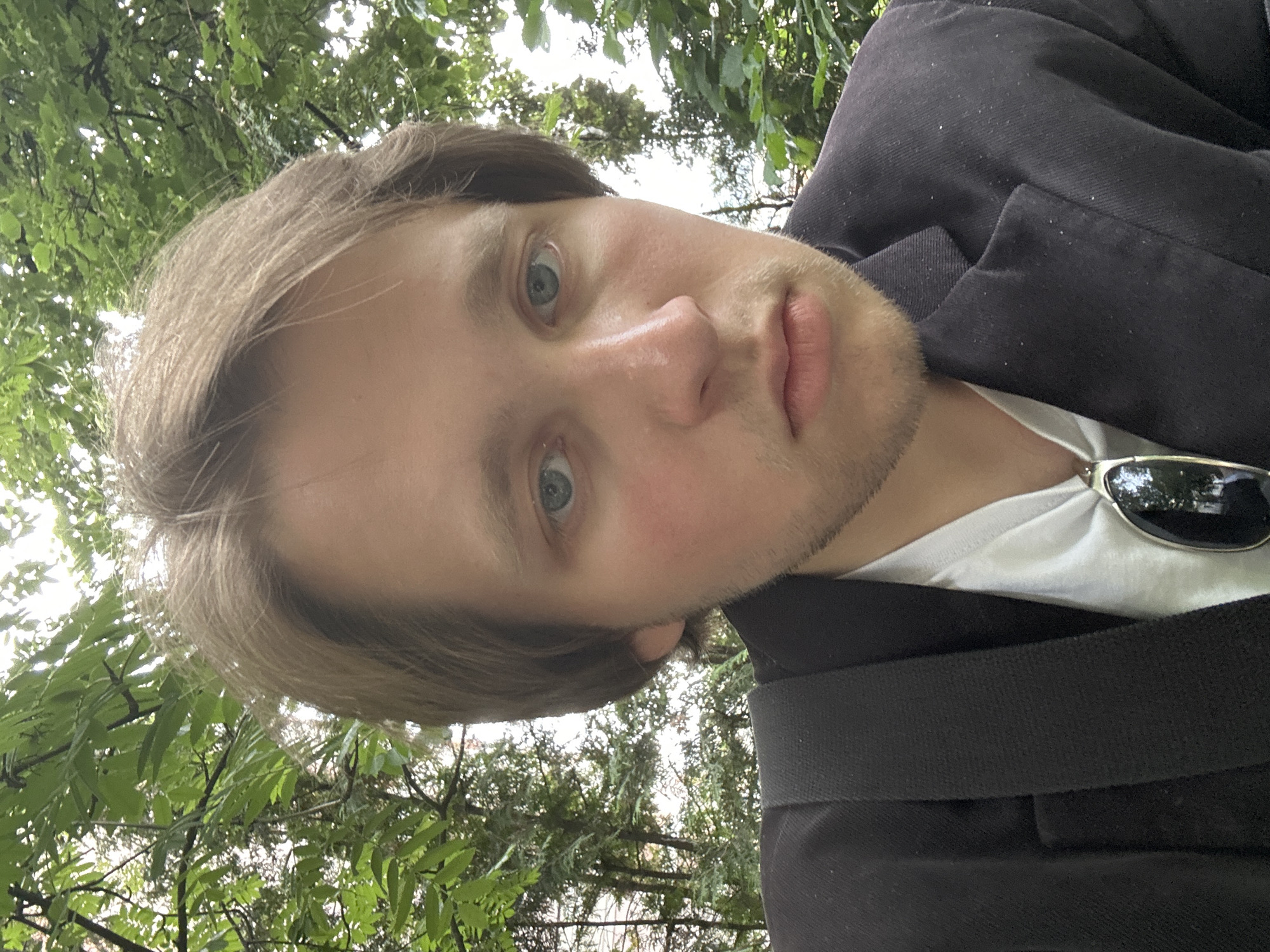 The image is a close-up, low-angle, selfie-style shot of a young person with light brown hair, blue eyes, and fair skin. They are wearing a black jacket over a white t-shirt, and a pair of dark sunglasses is visible on the jacket's collar. The person has a faint stubble on their chin and cheeks. Their expression is neutral, with their eyes looking directly at the camera.
The background is filled with lush green foliage, suggesting an outdoor setting, possibly a park or a wooded area. The lighting indicates it is daytime, with sunlight filtering through the leaves, creating bright spots and dappled shadows. The overall impression is of a casual, outdoor portrait. No text is visible in the image.