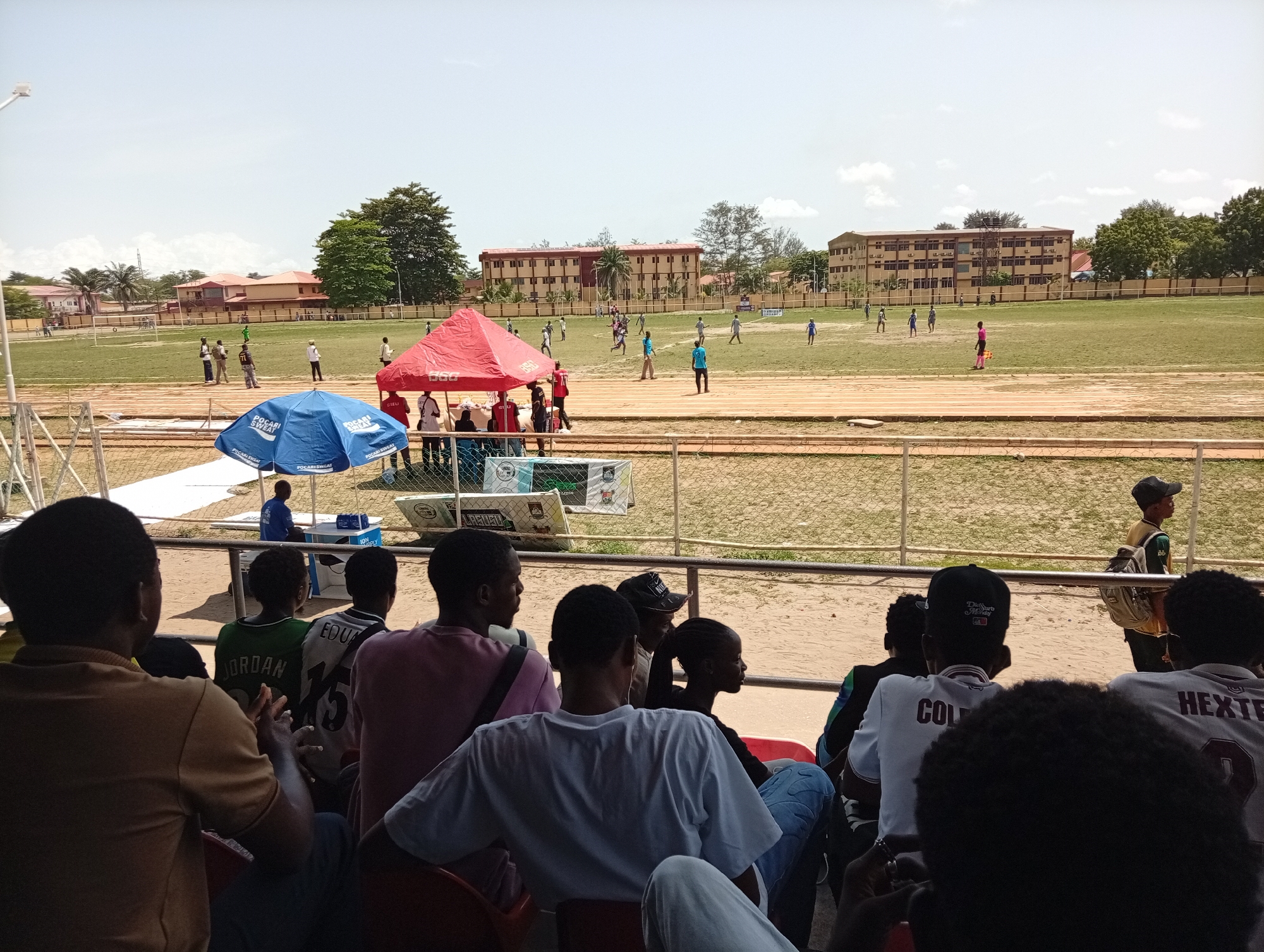 The image shows a soccer game being played in Lagos, Nigeria. Spectators sit in stadium seating behind a metal railing. A red tent and blue umbrella are positioned near the sidelines. A multi-story brick building can be seen behind the field. Players are actively engaged in the game, with several individuals spread across the field. A track surrounds the playing surface. The day appears bright with a mostly clear sky.