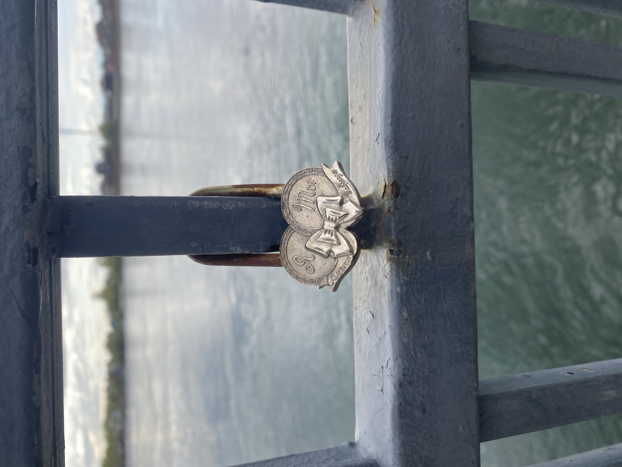 Here is a description of the image:
A close-up, rotated image shows two silver-colored coins or tokens attached to a dark gray metal structure. The background features a blurry view of water and indistinct land or buildings. The metal structure appears to be a railing. The coins are circular with raised designs and lettering in a cursive script, possibly in Russian. One of the tokens has a ribbon or banner element attached. The metal of the railing shows signs of wear, including peeling paint and rust. The photograph appears to have been taken during daylight. The location is Irkutsk, Russia.