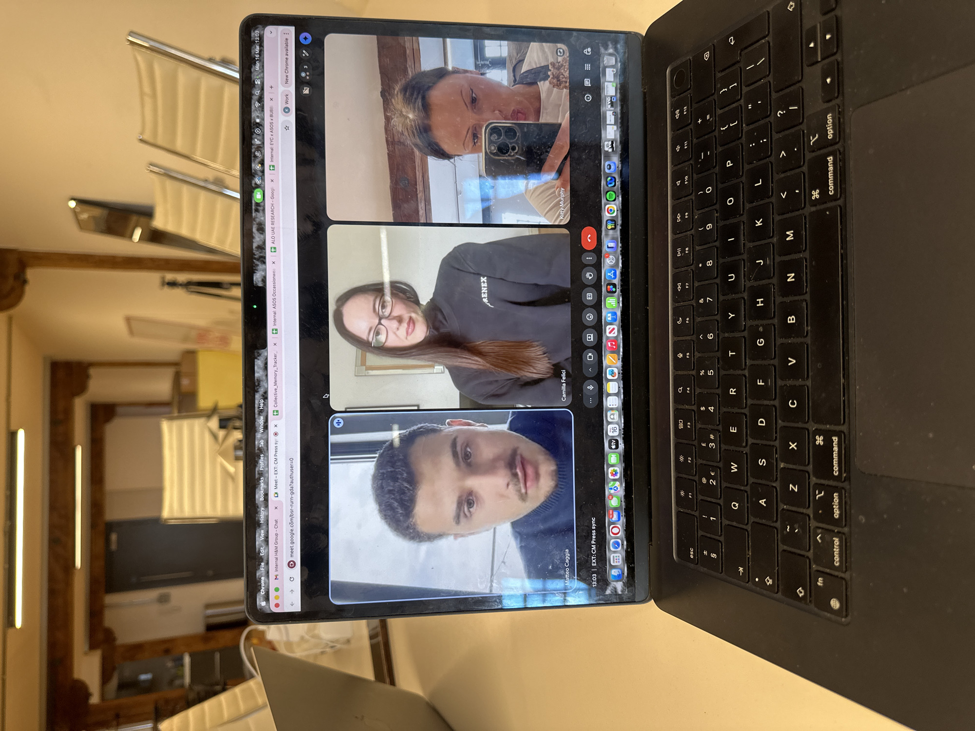 A silver MacBook Pro laptop is positioned on a light-colored surface within an interior space in the city of London, United Kingdom. The laptop's screen prominently displays a Google Meet video conference featuring three participants in a tiled layout.
The leftmost video panel shows a man identified as "Matteo Caggia", with short dark hair, a mustache, and stubble, wearing a dark blue turtleneck and looking directly forward. The central panel depicts a woman identified as "Camilla Felici", with long dark hair, wearing glasses, and a black sweatshirt with "RENEX" printed in white. The rightmost panel features a woman identified as "Kitty Murphy", whose face is partially obscured by a dark smartphone she is holding in front of herself, showing a reflection and a slight pouting expression.
The laptop's macOS interface is visible at the top, showing "Mon 16 Mar 13:03" and multiple Chrome browser tabs, including an active tab titled "Meet - EXT: CM Press sync". The bottom of the screen displays the macOS Dock with numerous application icons. The physical black QWERTY keyboard shows minor dust accumulation.
The background behind the laptop reveals an indoor environment, likely an office or studio. Visible elements include exposed wooden ceiling beams and two linear fluorescent light fixtures overhead. A dark grey door is centered in the background, and to the right, several contemporary office chairs with light-colored seats and chrome frames are arranged. A camera tripod and other indistinct objects are also discernible. The walls are predominantly light in color.