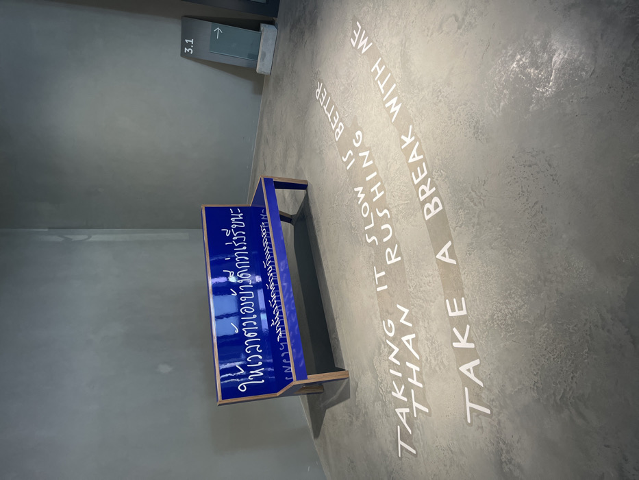 The image depicts an interior view within the Bangkok Art and Culture Centre (BACC) in Bangkok, Thailand. The scene features a minimalist architectural style with textured grey concrete walls and a light grey concrete floor. A glossy royal blue bench with natural wood-colored legs is positioned against the back wall, slightly off-center to the left. White Thai script is inscribed on the backrest of the bench, translating to "Give yourself some time, it's better than rushing." Directly in front of the bench, on the concrete floor, white stenciled English text reads: "TAKING IT SLOW IS BETTER THAN RUSHING" and "TAKE A BREAK WITH ME." In the upper right corner of the frame, a dark grey rectangular directional sign displays "3.1" and a white arrow pointing right, with a clear or frosted glass panel beneath it. The space is unoccupied by people, and the lighting is even and artificial.