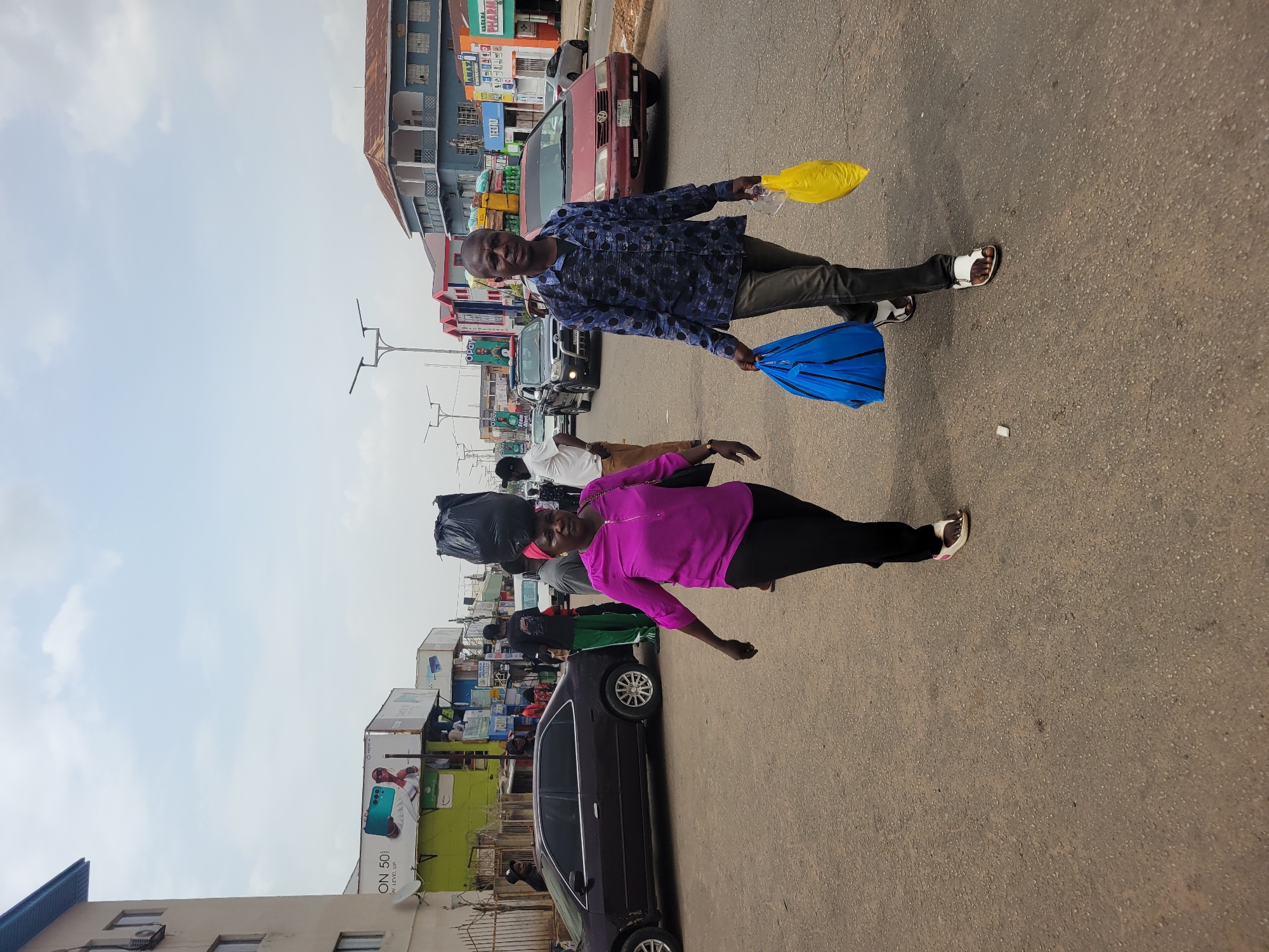 Two individuals are seen walking on a street, one man and one woman. The man is holding a yellow bag in his right hand and a blue bag in his left, while the woman has a large black bag balanced on her head. Behind them, there are several vehicles parked, and various commercial buildings line the street. Billboards and advertisements are also visible. The location appears to be an urban area, likely in Jos, Nigeria.