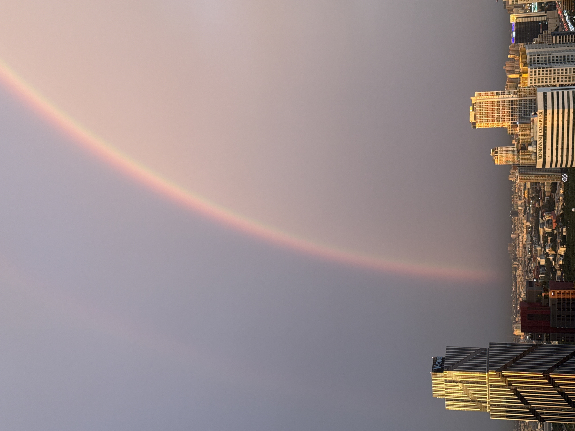 This media file depicts a panoramic view of a cityscape at dusk, featuring a prominent rainbow arching across the sky. The scene is dominated by tall, modern skyscrapers, characteristic of a dense urban environment. One building in the foreground displays the text "VONGVANIJ COMPLEX" on its facade, indicating a specific location within Bangkok, Thailand. The sky is a soft gradient of pale pink and gray, suggesting either sunset or the aftermath of rain, a common condition for rainbow formation. The lighting is dim, with a golden hue reflecting off the windows of some buildings, hinting at the last rays of sunlight. The overall mood is serene and awe-inspiring, with the natural phenomenon of the rainbow contrasting beautifully with the man-made structures of the city. The image captures a moment of natural beauty over a bustling metropolitan area.
