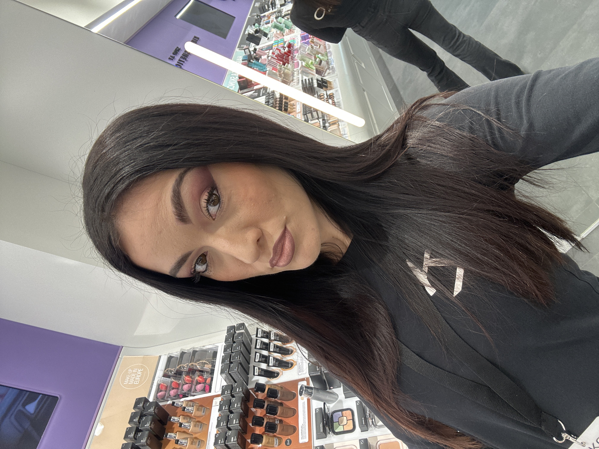A woman with long, dark hair, dark eyebrows, and brown eyes takes a selfie in what appears to be a makeup store. The image is rotated, showing her head tilted as she looks directly at the camera. She wears pink eyeshadow and a muted pink lipstick. She is dressed in a black long-sleeved top with a white/silver logo (resembling a stylized 'K') on the chest and a black lanyard.
The background reveals a modern retail environment with white and purple shelving units displaying various cosmetic products, including foundation bottles, compacts, and beauty sponges. Bright fluorescent lights illuminate the space. A large reflective surface, likely a mirror, shows the reflection of her lower body (wearing dark pants) and other parts of the store.
Visible text includes a circular label stating "MAKEUP MADE IN EUROPE" and partially visible, rotated text on a display sign, which seems to include "PUBLIC" and "24/S TI". The woman has a neutral to slightly smiling expression.