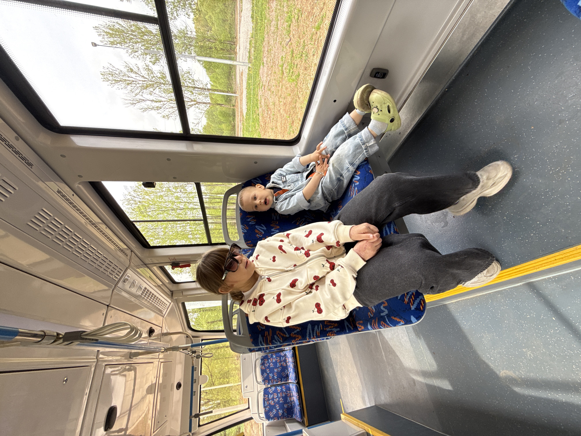 A person wearing a white jacket with red cherries and sunglasses is sitting in a blue-patterned seat on a bus, with a young child wearing a denim jacket and light-green shoes laying in their lap. The child has their feet on the wall of the bus. The bus has a large window showing greenery. Other seats are visible in the background. The location appears to be a bus travelling through a city.