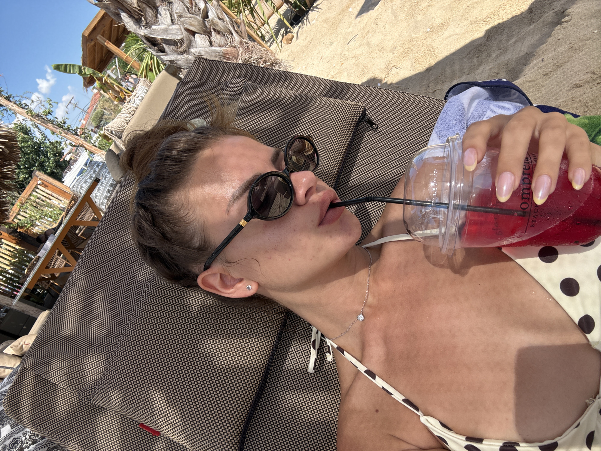 A young woman with dark hair is lying on a sun lounger on a sandy beach, wearing a polka dot bikini and sunglasses. She is holding a plastic cup with a red drink and a straw. Behind her, there are tables and chairs, palm trees, and buildings, suggesting a beachside cafe or resort. The sky is clear and blue.