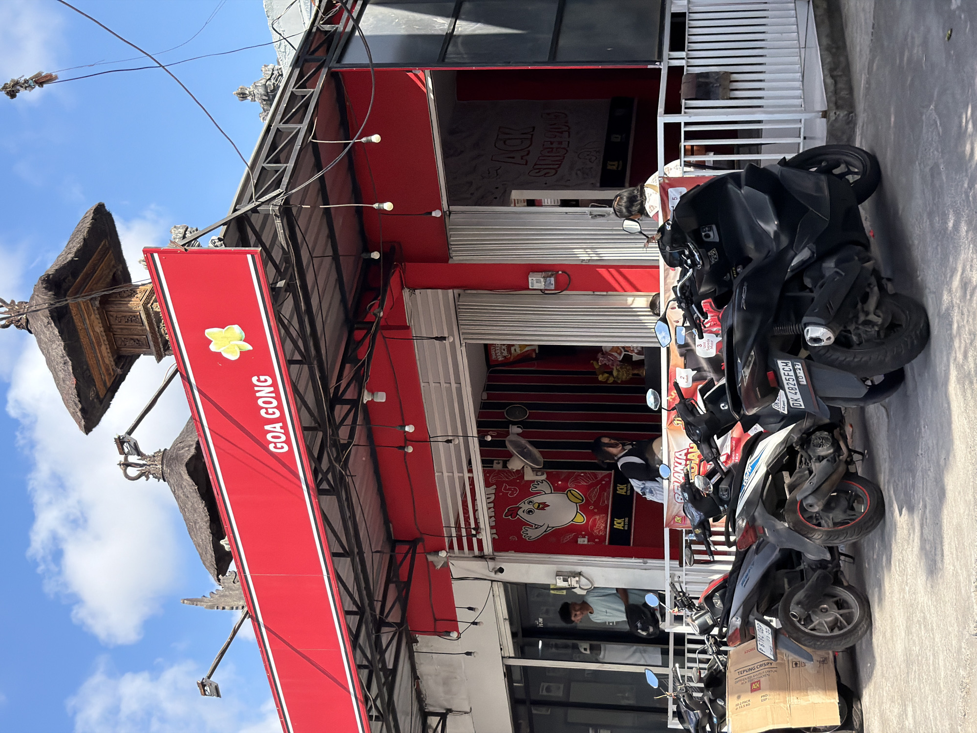 The image depicts a daytime street scene in Kuta Selatan, Indonesia, under a bright blue sky with scattered white clouds. The central subject is a commercial building with a red and white facade, suggesting it's a food establishment, likely a fried chicken restaurant given the prominent chicken logo.
A large, vertically oriented red banner on the left side of the building displays the text "GOA GONG" in white capital letters, accompanied by a yellow plumeria flower graphic. Above this banner, traditional Balinese-style roof structures are visible, indicating the local architectural influence.
Inside the building, a wall is decorated with a large cartoon chicken mascot and the brand name "ACK" repeated multiple times, including "ACK SINCE 2015". Other partially visible text includes "SPENICK" or "SPICY K". String lights are strung under an awning above the entrance. The storefront features metal roll-down shutters, some partially open.
Several motorbikes are parked on the sidewalk in front of the building. One black motorbike in the foreground has a visible license plate: "DK 4825FCM" with "10-27". Another motorbike carries a cardboard box labeled "TEPUNG CRISPY" and "ACK", with a registration number "P 6784 DBP" and date "03-30".
Two people are present: one person is partially seen near a motorbike, wearing a dark and white top, and another individual is visible through a glass door or window inside the building, wearing a light blue shirt. The bright sunlight indicates it is daytime.