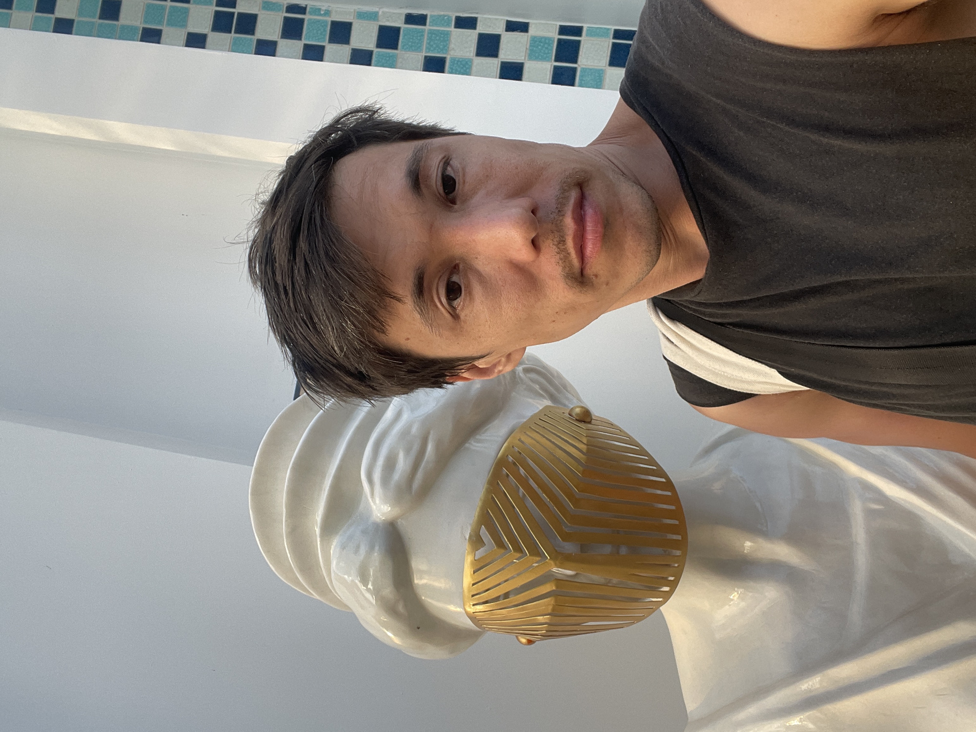 A man stands beside a white, glossy statue of a woman's head, the statue's face partially obscured by a geometric, golden-colored mask. The man wears a black t-shirt with white trim. The background includes white walls and a border of blue and white tiled squares. The location is presumably in Nha Trang Ward, Vietnam.