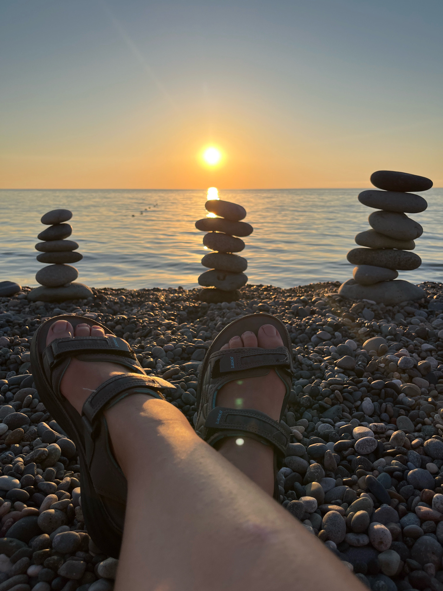 The image depicts a close-up view of a person's legs and feet, clad in dark gray sandals, resting on a pebbled beach. The perspective is from the person's point of view, looking out towards the sea. In the mid-ground, three towers of stacked stones are arranged in a line. The sun is setting over the calm sea, casting a warm glow across the scene and illuminating the sky with shades of orange and yellow. The beach is covered in various sizes and colors of smooth pebbles. The background features the tranquil sea meeting the horizon, under a gradient sky that transitions from a pale blue to a warm orange hue. The scene is set at a beach in Kobuleti, Georgia, implied by the presence of the Black Sea and the characteristic pebbled shore.