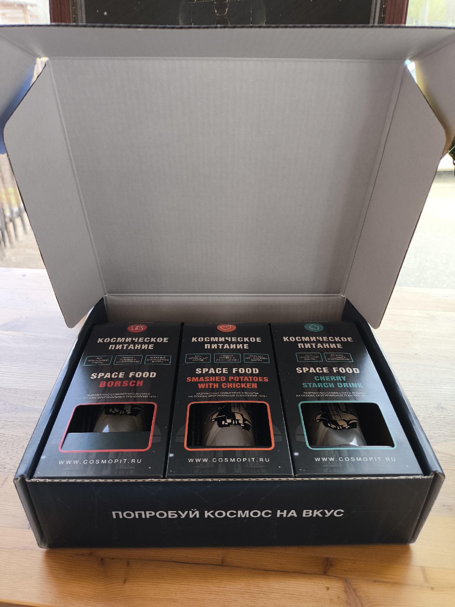 An open cardboard box, likely part of an unboxing, is seen from a slightly elevated angle, revealing three packages of "space food" from the brand "COSMOPIT.RU". The box rests on a light wooden surface, with a blurred background showing an outdoor area through a window, indicating a daytime setting.
Inside the box, three black packages are neatly arranged. Each package is labeled in both Russian and English as "SPACE FOOD." From left to right, the products are identified as "BORSCH" (Borscht), "SMASHED POTATOES WITH CHICKEN," and "CHERRY STARCH DRINK." Each package features a small transparent window that provides a glimpse of the product's tube-like container. The labels on all packages also state "КОСМИЧЕСКОЕ ПИТАНИЕ" (SPACE FOOD), "100% НАТУРАЛЬНЫЙ СОСТАВ" (100% NATURAL COMPOSITION), and "ПРОДУКТ ГОТОВЫЙ К УПОТРЕБЛЕНИЮ" (READY-TO-EAT PRODUCT). Additional text on each package indicates they were "РАЗРАБОТАНО СОВМЕСТНО С ВНИИТЭК НА ОСНОВЕ ОРИГИНАЛЬНЫХ ТЕХНОЛОГИЙ 1978 г." (DEVELOPED JOINTLY WITH VNIITEK BASED ON ORIGINAL TECHNOLOGIES OF 1978). The website "WWW.COSMOPIT.RU" is visible on the bottom of each package's front label.
The inside bottom flap of the box prominently displays the Russian slogan "ПОПРОБУЙ КОСМОС НА ВКУС," which translates to "TASTE SPACE." The overall presentation suggests a unique and possibly novelty culinary experience inspired by astronaut rations.