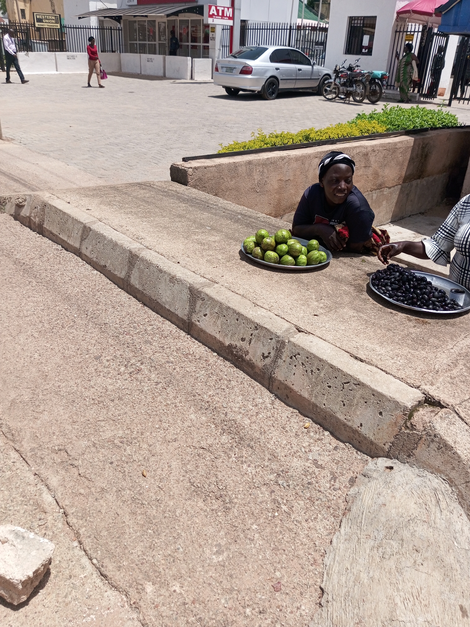 The image shows a street scene in Bukuru, Nigeria. Two women are visible in the foreground, sitting on a raised concrete area. One woman, wearing a dark t-shirt, is looking towards the right and smiling. She is leaning on a platter of green fruits. To her right, another woman, wearing a checkered shirt, is reaching for a platter of dark, oval-shaped fruits, likely olives. In the background, a silver sedan is parked, and two motorcycles are visible next to it. A building with "ATM" and "WESTERN UNION MONEY TRANSFER" signs is also present. The ground is paved with interlocking bricks. The lighting suggests it is daytime, with clear skies. The overall atmosphere appears to be a commercial area with informal street vending.