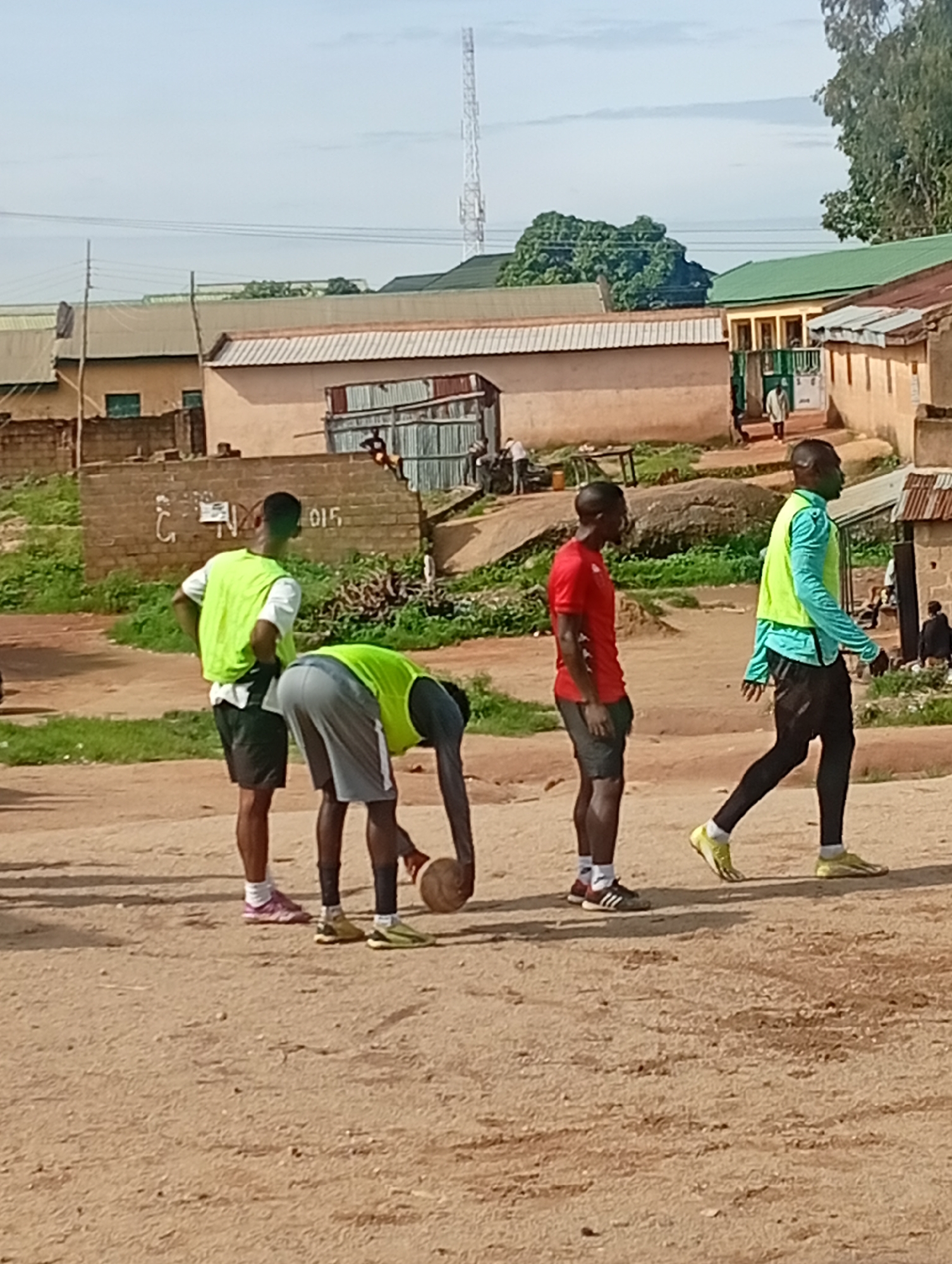 This image depicts an outdoor scene in Jos, Nigeria, under a clear, bright sky. Four young men are the primary subjects, engaged in what appears to be a casual sports activity.
In the foreground, two men are positioned close to each other. One man, wearing a bright lime green vest over a white t-shirt and black shorts, stands with his hands on his hips, looking towards the right. The other man, in a lime green vest and grey shorts, is bent over, interacting with a ball at his feet. His posture suggests he is either about to kick, pick up, or is otherwise in possession of the ball.
Further to the right, a man in a red t-shirt and black shorts stands facing left, looking towards the other individuals. To his right, a fourth man, also in a lime green vest and black leggings or long shorts, is walking to the right. His body is angled slightly away from the viewer, and he appears to be moving with purpose. His footwear includes yellow athletic shoes.
The environment is characterized by a dirt or sandy ground, with scattered patches of dry grass and some larger rocks or mounds of earth visible in the mid-ground. In the background, there are several buildings, including structures with corrugated metal roofs, brick walls, and some with painted facades. One building has a green roof and a series of windows. A tall communication tower is visible in the far background, a common landmark in urban or semi-urban areas. Power lines stretch across the upper portion of the image. The overall impression is of a slightly unkempt, utilitarian urban setting, possibly a community space or training ground.
