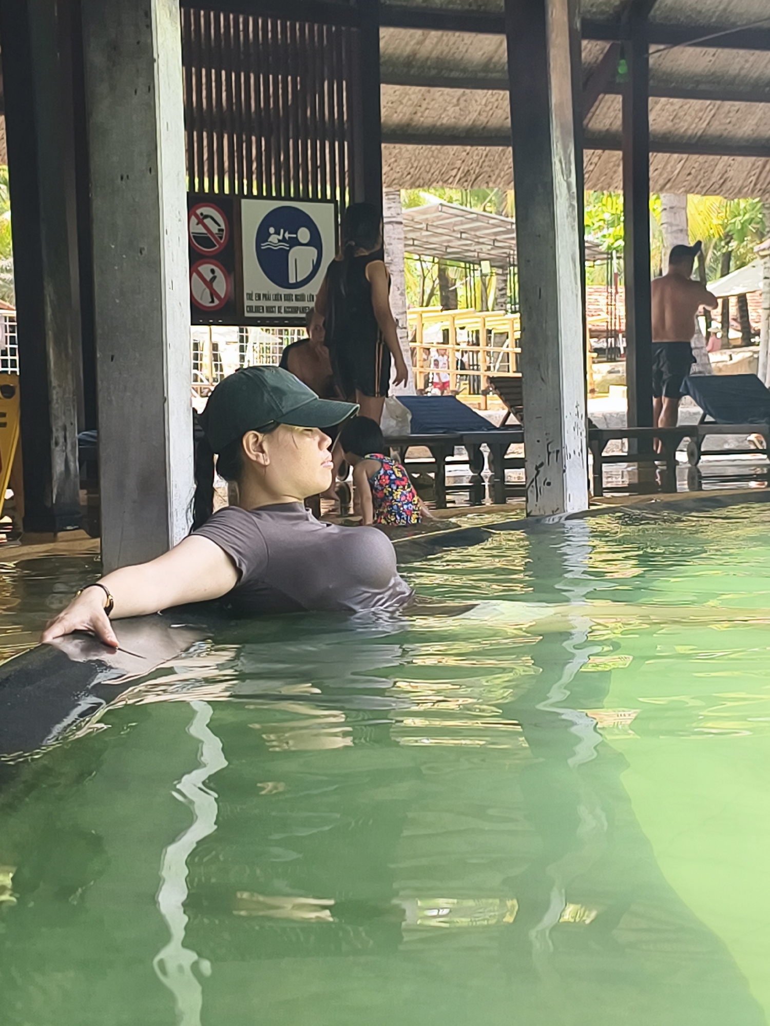 The image shows a woman in a body of water, possibly a hot spring or pool. She is wearing a dark-colored shirt and a dark baseball cap, and she is resting her arm on the edge of the pool. The water is green and murky, with reflections of light and objects on the surface. Behind her, there are other people in the area, including a man without a shirt and a child playing. There are wooden structures and beams surrounding the pool area, and signs with pictograms are posted on some of the beams. Lounge chairs and other beach furniture are visible in the background. The location is Unknown, Unknown.