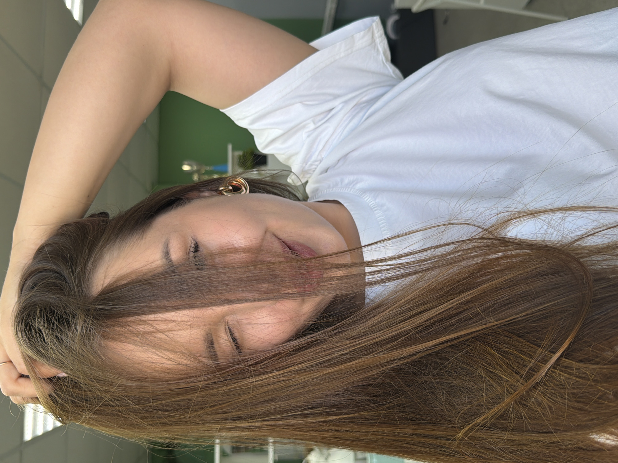 A young woman with long, light brown hair is posing in what appears to be an indoor setting. The woman is wearing a white t-shirt, and her right arm is raised, with her hand touching her hair. The lighting is bright. Background details are minimal, but suggest a modern interior.
The city of Chaikovsky, Russia, is not definitively present in the image, but it is possible that the scene is located there.