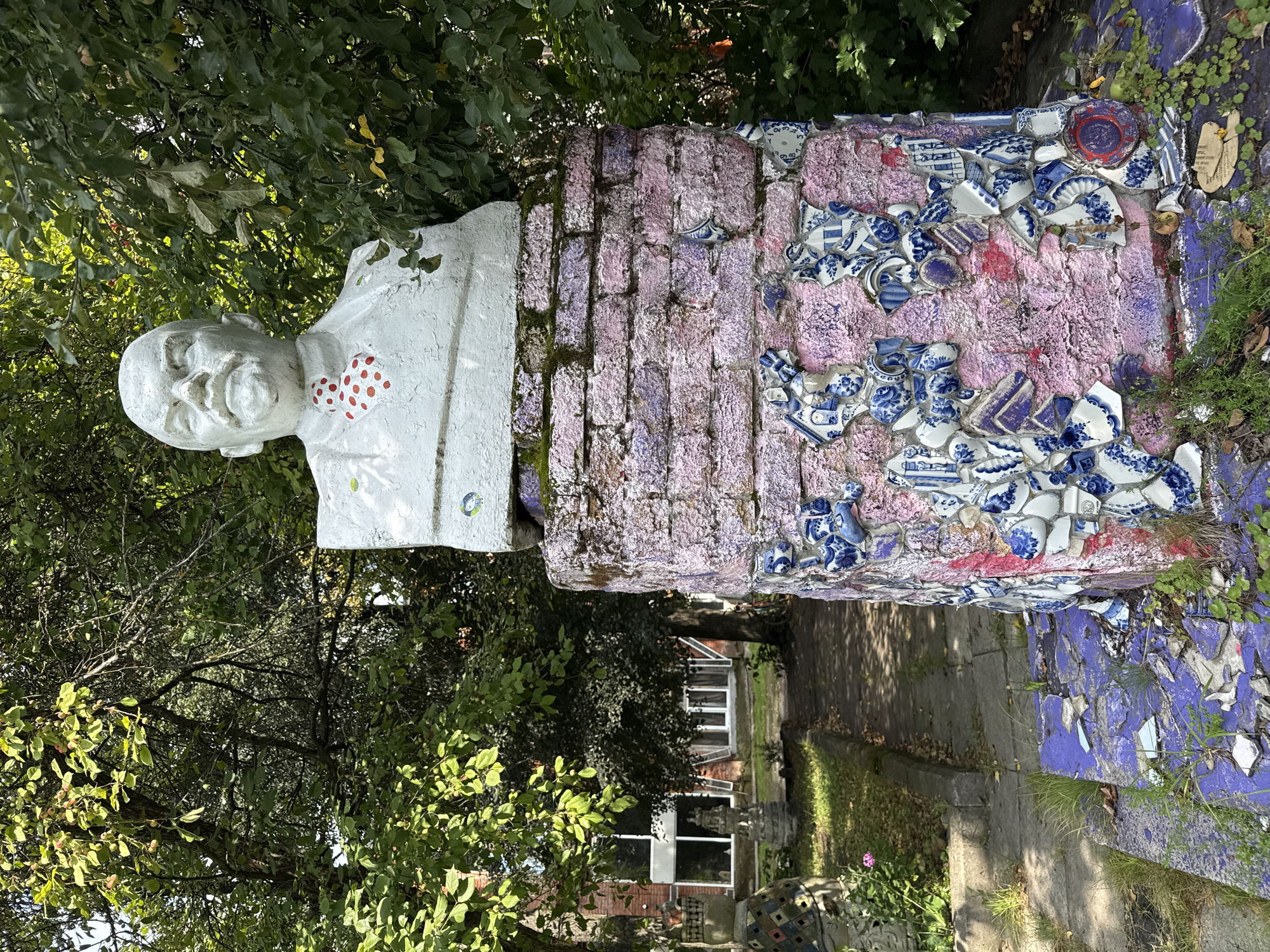 A bust of Vladimir Lenin, painted white with a red polka-dot tie, stands atop a brick pedestal that is adorned with broken blue and white ceramic tiles and pink and purple paint. The pedestal is surrounded by overgrown grass and some broken purple tiles. In the background, trees with green leaves provide shade, and a pathway leads to a building with large windows. The scene appears to be located outdoors, likely in a park or garden setting. The overall impression is one of decay and unconventional artistic expression.