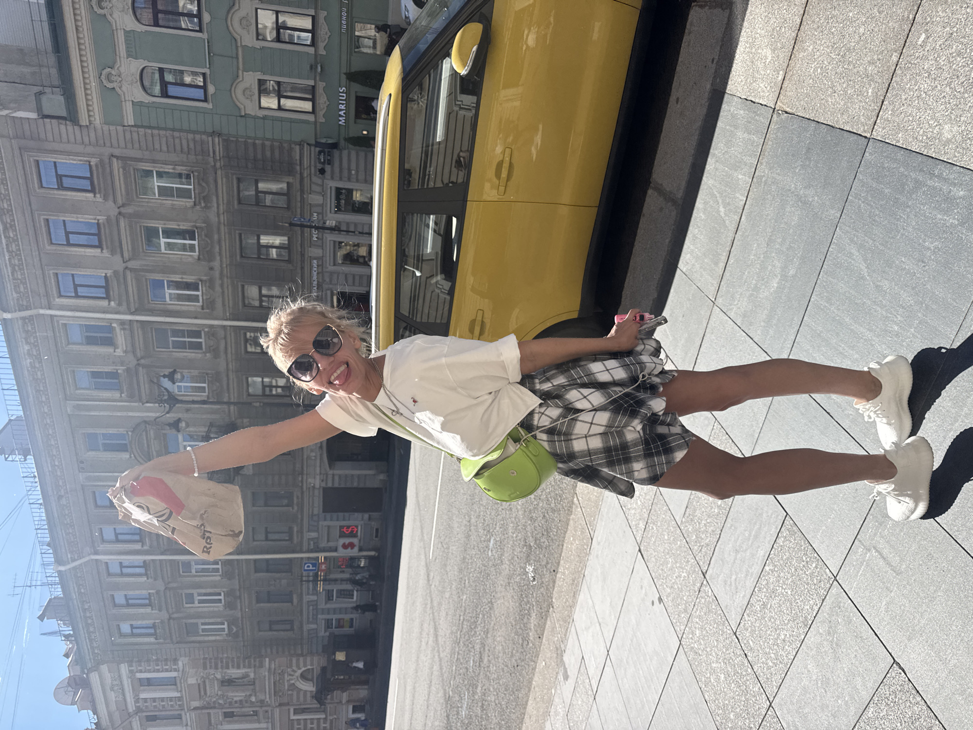A woman stands on a sidewalk in Saint Petersburg, Russia, holding a paper bag aloft with her right hand. She is wearing a white t-shirt, a black and white plaid skirt, and white sneakers. A bright green crossbody bag is slung over her shoulder. She is wearing sunglasses and has blonde hair. Behind her, a yellow car is parked on the street, and several historic buildings line the street. The sun is shining brightly, casting shadows on the sidewalk. The buildings appear to be residential or commercial, with multiple windows and ornate architectural details. A sign with the word "MARIUS" is visible on one of the buildings.