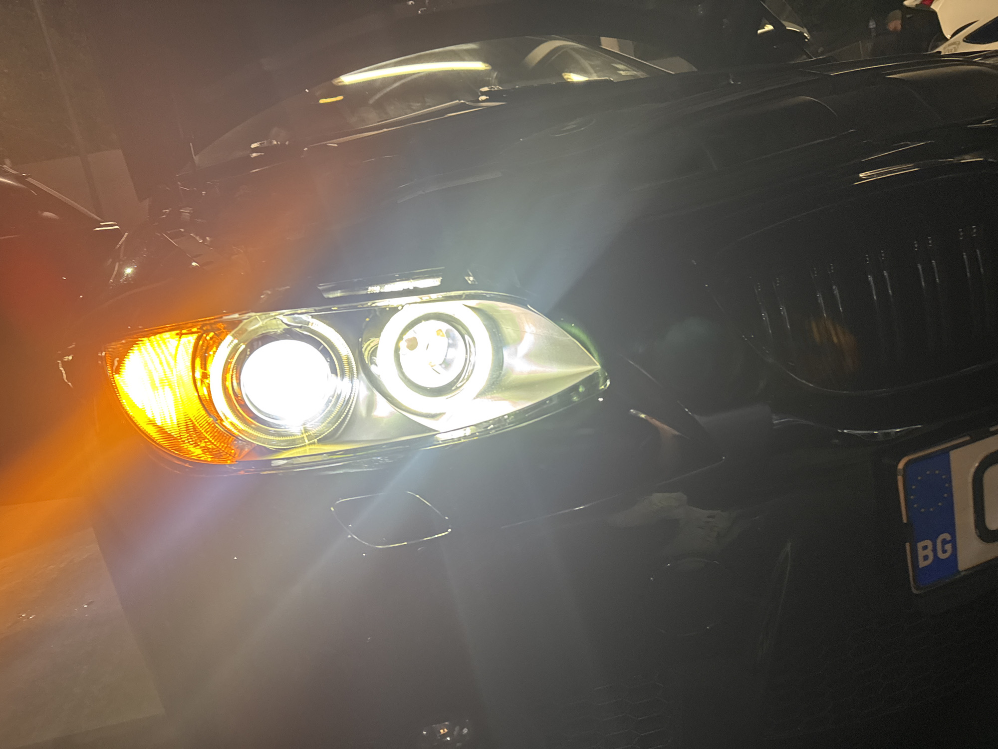 A close-up, low-angle shot captures the front end of a black BMW car in dark, low-light conditions, likely at night or indoors. The car's headlights are prominently illuminated, casting a bright glow. The main headlight assembly features two distinct white "angel eye" rings, with a bright white projector beam in the inner ring. To the left, the orange turn signal is also brightly lit.
The car's dark, vertical slat kidney grilles are visible on the right side of the frame. Reflections of overhead light sources are seen on the car's hood and windshield, suggesting an indoor setting or an area with overhead lighting, possibly a car wash bay. In the lower right corner, a portion of a Bulgarian license plate is visible, showing the blue EU strip with stars and the "BG" country code in white lettering on a blue background.
A faint figure of a person appears in the upper right background, seemingly looking towards the car. The surface beneath the car is dimly lit. The overall impression is a focused shot on the car's striking front lights, highlighting its design in a dark environment.