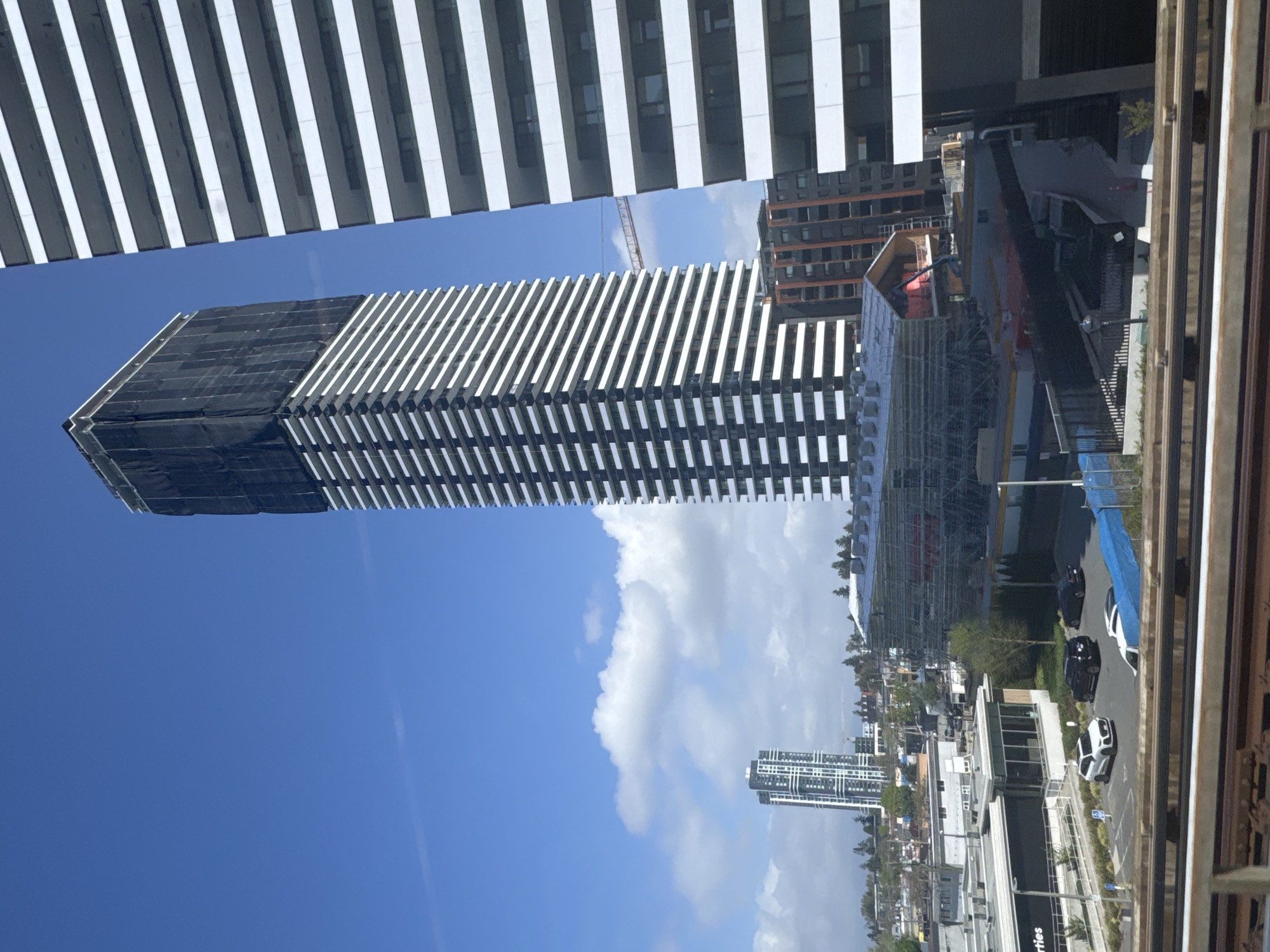 This image depicts a sunny daytime urban landscape in Surrey, Canada, dominated by modern architecture and ongoing construction.
The central subject is a tall, slender skyscraper under construction, featuring a distinctive design of horizontal white fins along its facade. The upper portion of this building is covered in black protective netting. Framing the top of the image is a section of another contemporary building, also characterized by similar white horizontal elements.
In the middle ground, a construction crane is visible against the clear blue sky, which has scattered white clouds. The surrounding area includes other buildings, some appearing residential or commercial, extending into the distance.
At ground level, a street or parking area is visible with several parked cars, including a white vehicle resembling a Porsche and a couple of dark sedans. Blue tarps, possibly construction barriers, are also present. Scaffolding and other construction materials are noticeable around the base of the main skyscraper and adjacent structures. Part of the word "rties," likely from "Properties," is visible on a building at street level. The overall scene suggests a busy, developing urban environment.