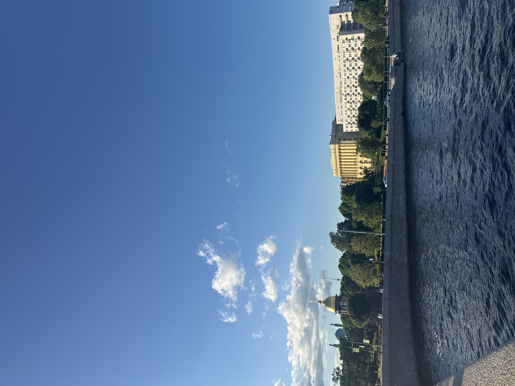 The image captures a waterfront view of Saint Petersburg, Russia, on a clear day with a bright blue sky and scattered clouds. In the foreground, a dark blue river with ripples stretches across the bottom of the frame. A stone embankment lines the river, with a paved walkway on top.
Beyond the embankment, a line of lush green trees creates a natural barrier between the walkway and the buildings. To the left of the trees, a prominent golden dome, likely part of a church or cathedral, stands out against the sky, with a cross on top. To the right of the golden dome, two other smaller domes are visible.
Further along the embankment, a light yellow building with classical columns and a light-colored modern apartment building with many windows dominate the skyline. Several cars are visible on the street behind the trees, and a few pedestrians can be seen walking along the embankment. The overall impression is a serene urban landscape showcasing the city's architectural beauty alongside its natural elements.