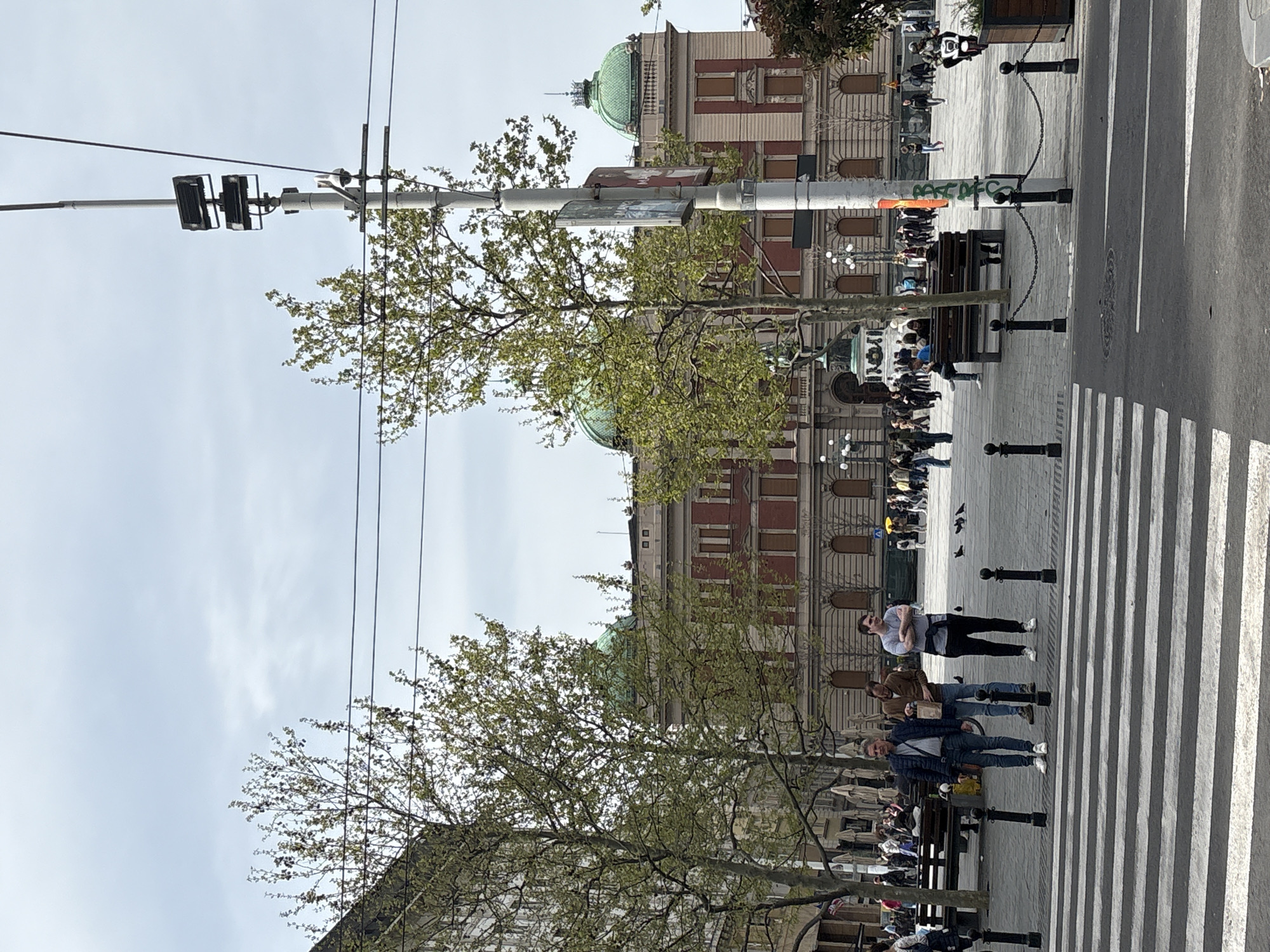 The image captures a daytime street scene in Belgrade, Serbia. In the background, there is a building with red brick and stone detailing. The building has a green dome.
A pedestrian crossing is visible in the foreground, with people walking across. Several individuals are sitting on benches. A tree with new spring foliage is between the camera and the building. There is also a utility pole with wires and what appear to be pedestrian signals attached.