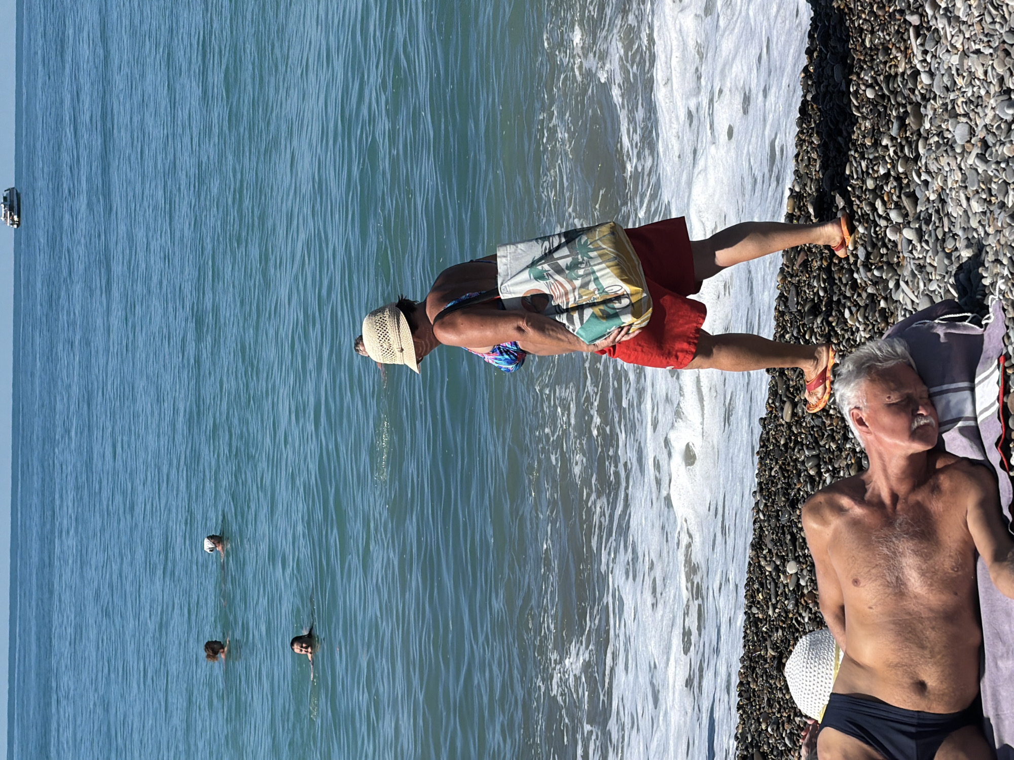 The image depicts a sunny day at a pebble beach in Sochi, Russia. In the foreground, an elderly man with grey hair and a mustache is sleeping on his back on a towel, wearing black swimming trunks. Next to him is a white straw hat. A woman wearing a straw hat, a floral bikini top, and red shorts is walking on the beach, carrying a patterned tote bag over her shoulder. She is walking towards the right of the frame. In the background, several people are swimming in the blue, rippling sea. Further out in the water, a white boat with a superstructure is visible on the horizon under a clear blue sky. The waves are gently breaking on the pebble shore.