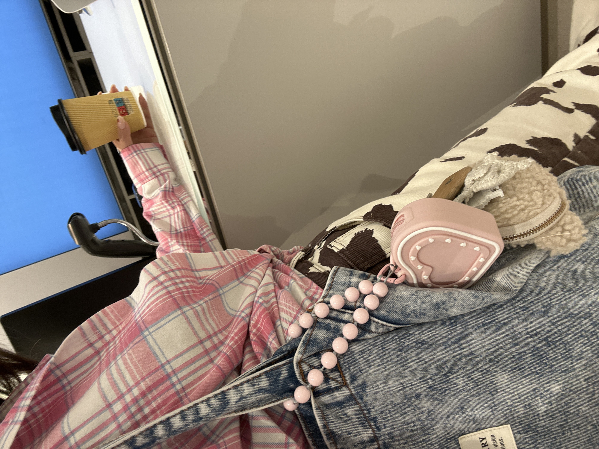 The image shows a person indoors, captured from a tilted, overhead perspective. The person's right arm holds a yellow, corrugated paper cup with a black lid, which has a partially visible red and blue logo or emblem on its side. They are wearing a pink, white, and light blue plaid long-sleeved shirt, layered with a light blue denim garment, possibly overalls or a jacket. A string of shiny pink beads is draped across the denim.
Resting on the denim and a brown and white cow-print fabric (likely a blanket or pillow) is a small, pink, heart-shaped item, possibly an AirPods case or a small pouch, adorned with multiple tiny white heart outlines. Adjacent to this is a fluffy, light-colored object, which may be a bag charm, with a small wooden attachment. In the background, a portion of a blue screen, likely from a monitor or television, and part of a black gooseneck lamp are visible. A fabric tag on the denim garment displays partially legible text including "RY", "vision", and "tions.". The scene suggests a relaxed, casual moment indoors. The location of Šušanj, Montenegro, is not indicated by any visual cues within the image.