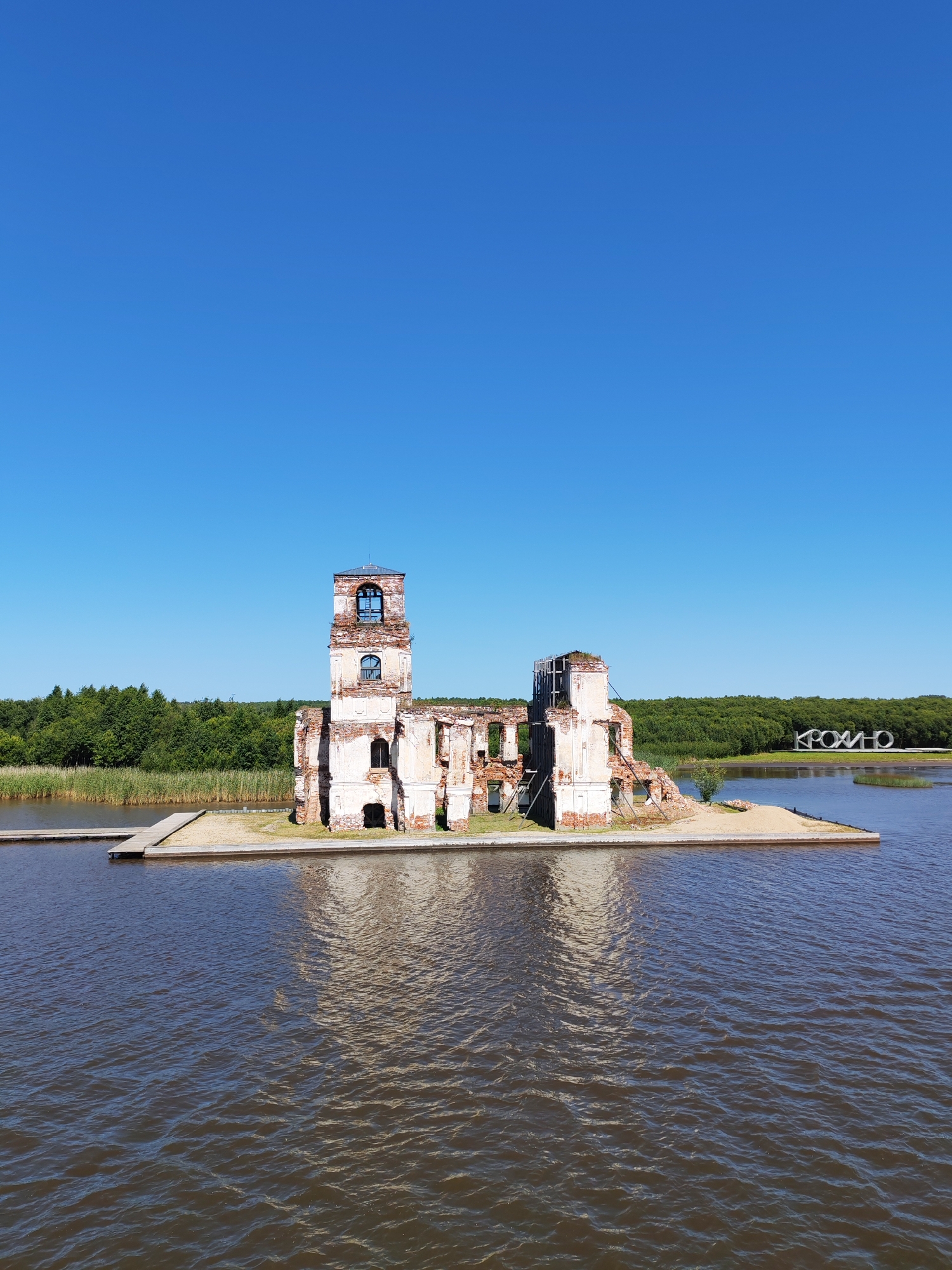 The image captures a sunny day in Krokhino, Russia, featuring the ruins of a former church. The structure, made of brick and whitewashed stone, stands on a small island surrounded by dark, rippling water. A wooden pier extends from the left side of the island towards the foreground. Behind the ruins, a dense green forest lines the horizon, with a clear blue sky above. In the distance, on the right side of the image, a large white sculpture spelling out "KROKHINO" is visible. There are no people or animals present in the scene.