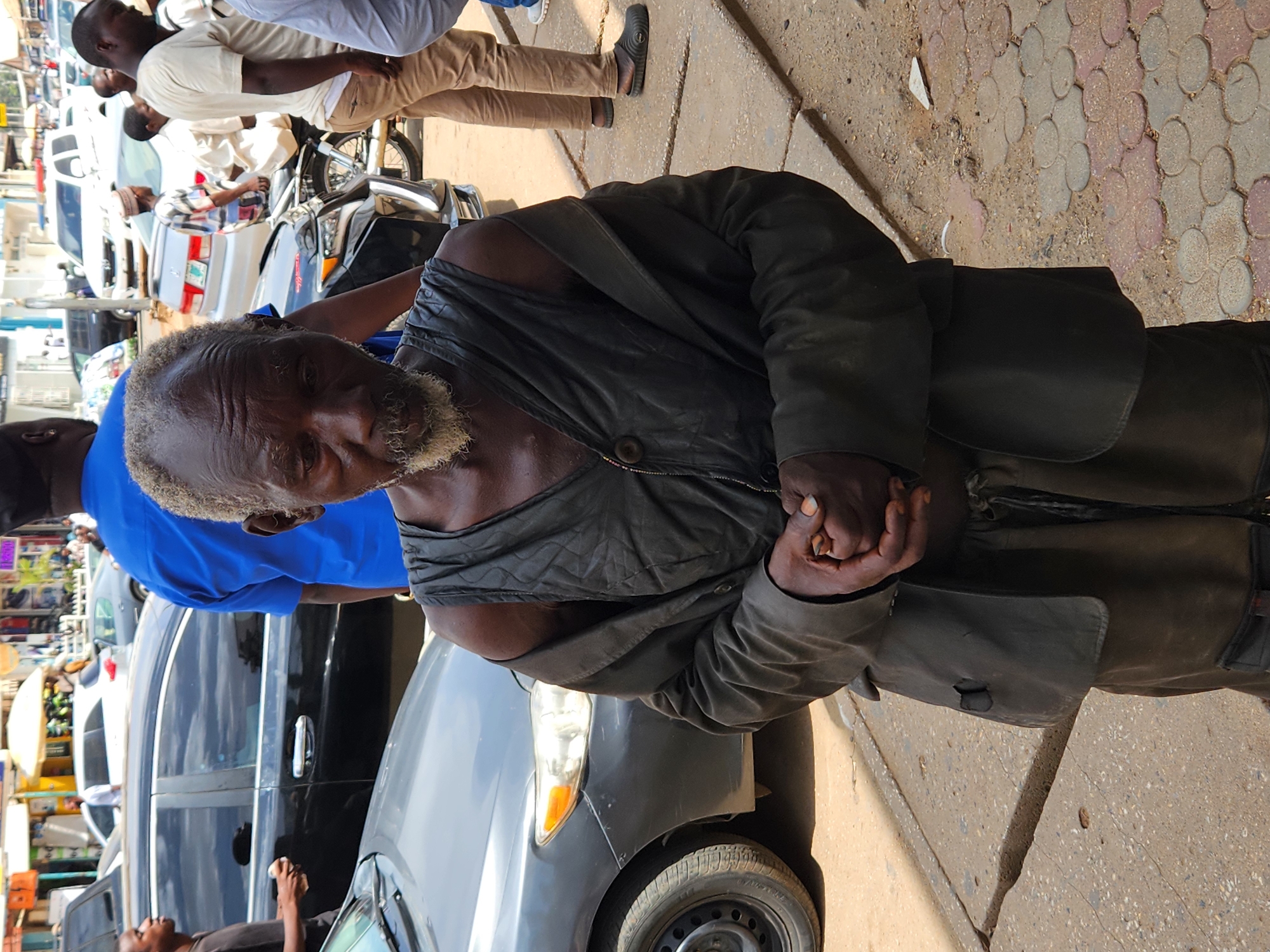 An eye-level, outdoor photograph shows an elderly man with a weathered face standing on a street. The man is wearing a dark, worn jacket and pants. His hair is graying, and he has a short, salt-and-pepper beard. He looks towards the camera, holding his hands cupped together in front of him.
Behind him are several parked cars, some with people standing nearby. A man in a blue shirt is standing partially in front of the car in the foreground, partially obscuring the view of the street beyond. Other people are milling about in the background, and there are buildings visible across the street. The ground is paved, and the lighting suggests it is daytime.
The location is Unknown, Unknown.
