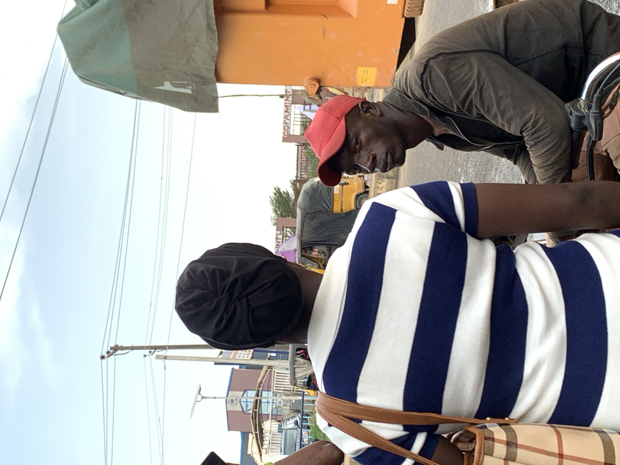 The image depicts a candid street scene in an urban area, likely Ikorodu, Nigeria, under an overcast sky. The photograph is taken from a perspective looking slightly upwards.
In the immediate foreground, seen from behind, is a person wearing a black head covering and a navy blue and white striped long-sleeved shirt. A brown shoulder bag with a plaid pattern is slung over their shoulder. Part of a smartphone is visible in their left hand.
In the middle ground, a dark-skinned man wearing a red baseball cap and a dark brown, possibly padded, jacket is leaning forward. He appears to be looking down towards the person in the foreground, possibly engaged in a conversation or an interaction involving a motorcycle or scooter, the handlebars of which are partially visible.
The background reveals typical street elements. On the right, a large orange container or the side of a truck is prominent. Behind the man in the red cap, a building displays the white text "COFAMINT". Further down the street, a yellow auto-rickshaw (keke napep) and other blurred vehicles and structures are visible, suggesting a bustling environment. Numerous power lines crisscross against the bright, cloudy sky, with utility poles supporting them. A green tarp or awning with a white logo, resembling a stylized bird or figure, hangs in the upper left corner of the frame.