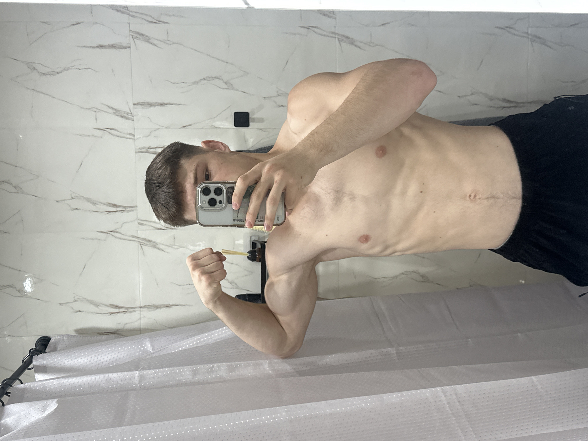 A young man with short brown hair is standing in front of a marble-tiled wall, likely in a bathroom, as evidenced by a white shower curtain on the left and a shelf with toiletries in the background. He is holding a smartphone in his right hand, angled to capture a selfie, and flexing his left bicep with his left hand. He is wearing black athletic shorts. The lighting appears to be artificial, possibly from overhead bathroom lights, creating soft shadows. The image is a close-up, focusing on the man's torso and upper body. There is no visual information to ascertain the location in Birobidzhan, Russia.