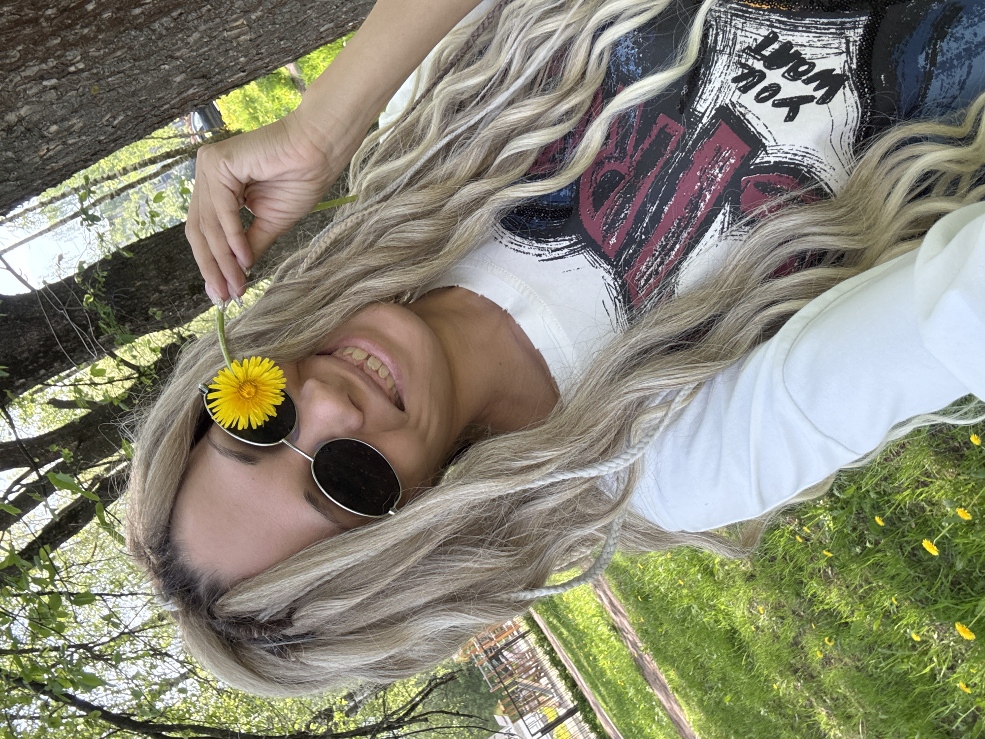 A fair-skinned woman with blonde braids lies on her back in a grassy area in the city of Rusanovo, Russia. She wears round, dark sunglasses. A yellow dandelion is positioned on one lens of the sunglasses. The woman smiles. She wears a white t-shirt with a graphic design. A tree trunk is visible to the left of the woman. Green grass and scattered yellow dandelions surround her.