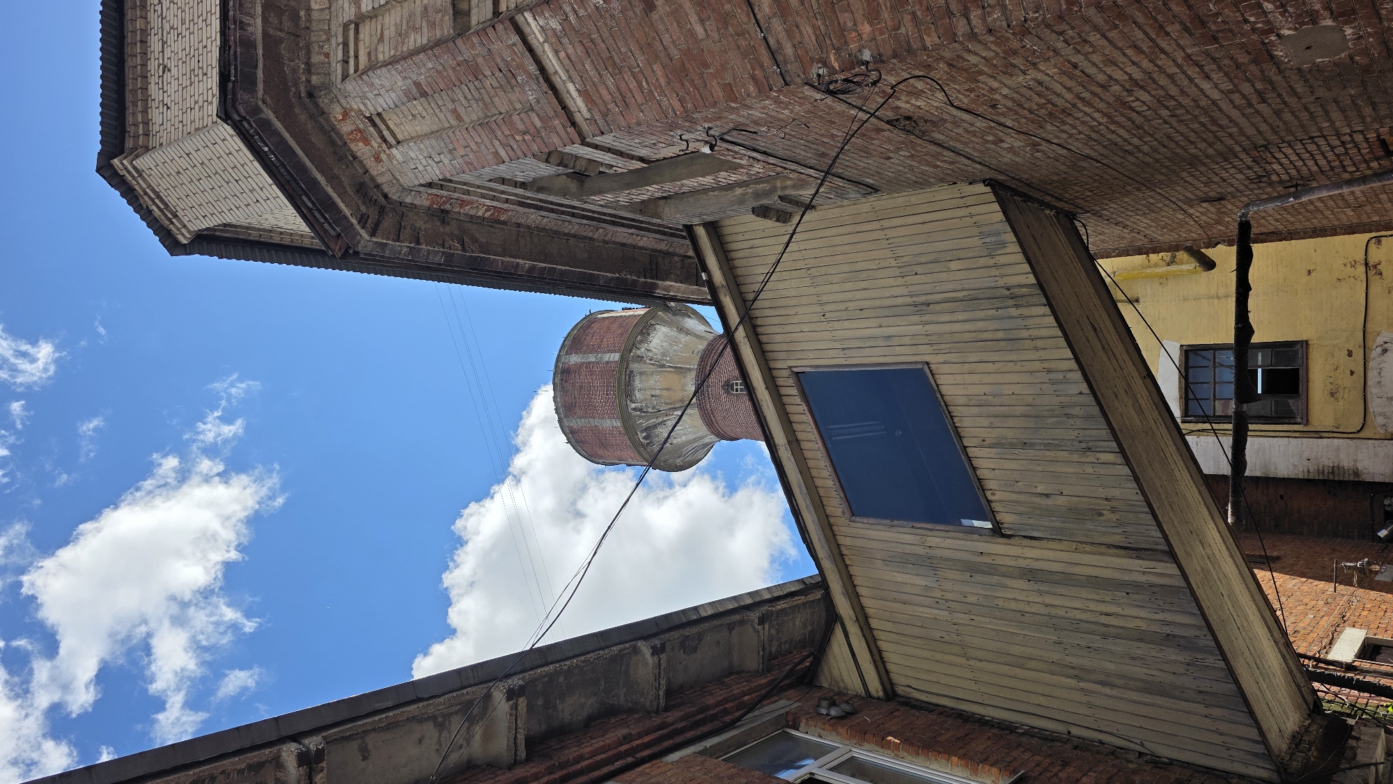 This low-angle, full-shot photograph captures a view of an old industrial area under a bright blue sky with scattered white clouds. In the center background, a red brick water tower with a concrete base stands prominently. In the foreground and mid-ground, the weathered facades of industrial buildings are visible. A wooden skybridge, likely connecting two structures, extends diagonally across the frame, featuring a large dark window. The buildings are constructed of red and beige brick, with some showing signs of age and disrepair. Power lines are visible crisscrossing the sky. The scene is located in Ivanteyevka, Russia, suggesting an urban industrial setting. There are no people or animals present.