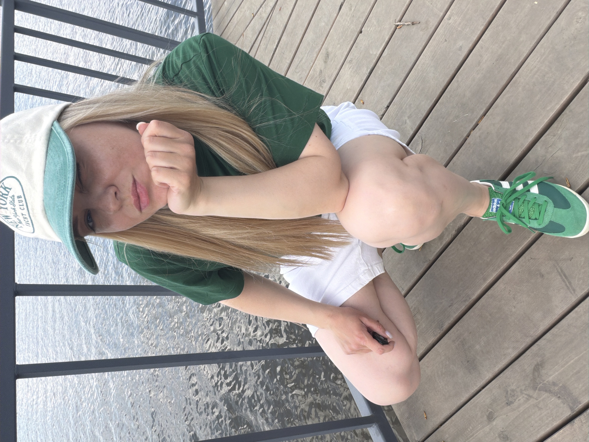 A young woman is crouching on a wooden deck near a body of water, likely a lake or river. She is wearing a green t-shirt, white shorts, and green Adidas sneakers. A baseball cap with the words "New York Yacht Club" is on her head. The background shows a metallic railing, and beyond it, the water. The location is not specifically discernable as being in the city of Votkinsk, Russia from the provided image, but we could infer she is near the Votkinsk Reservoir.