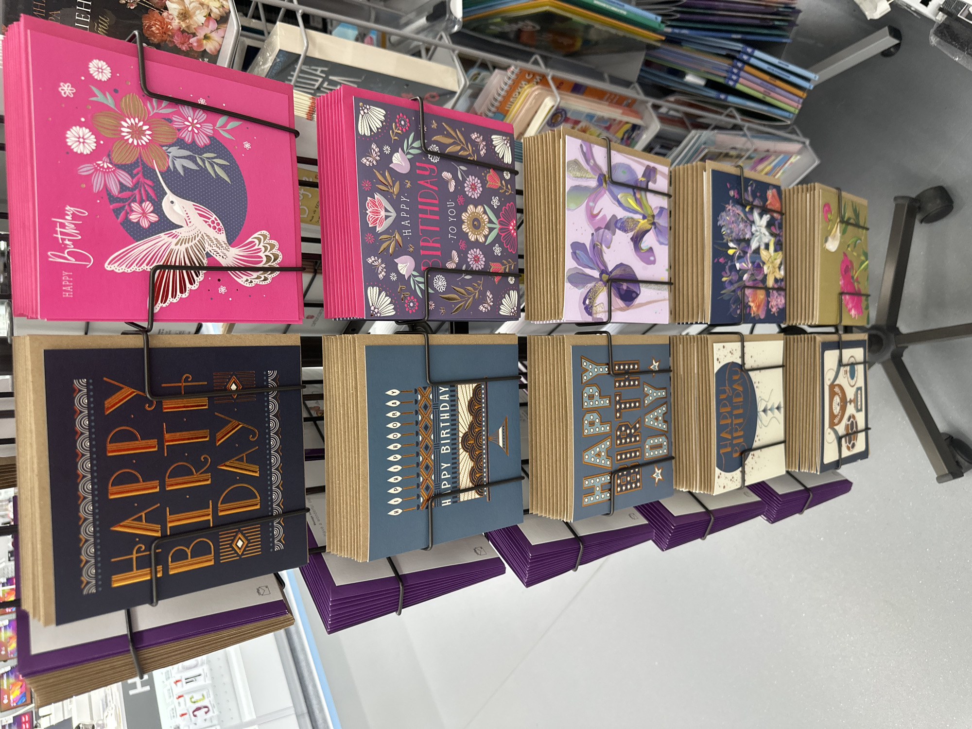 The image shows a display of greeting cards, likely for birthdays, in a retail setting. The cards are arranged in rows on a metal rack, with each design stacked and secured with a metal band. The cards feature various designs, including floral patterns, hummingbirds, and stylized "Happy Birthday" messages. The colors range from bright pink to muted blues and greens. The environment appears to be a store aisle with other merchandise visible in the background.