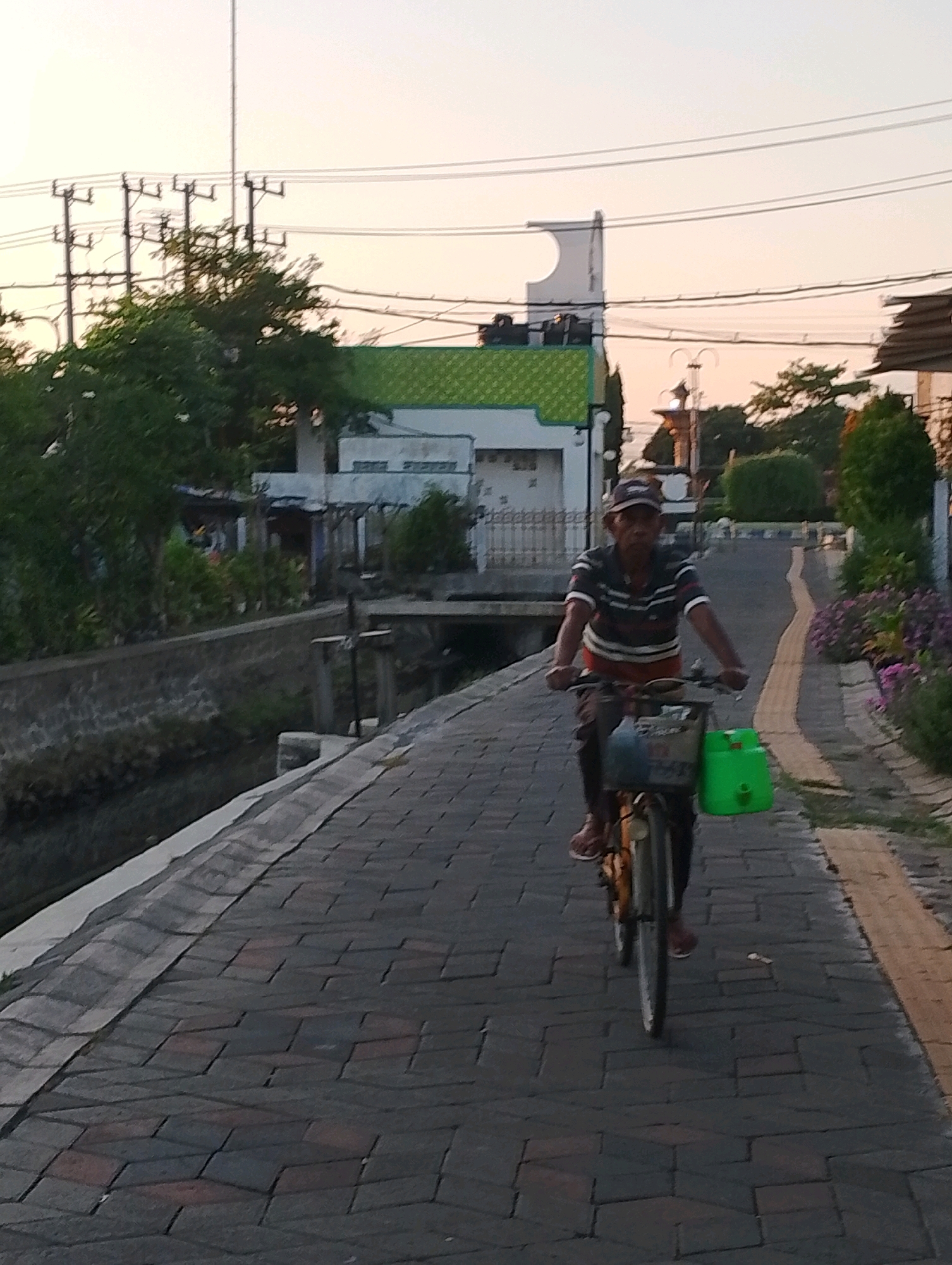 An elderly Indonesian man rides a bicycle on a paved street in Probolinggo, Indonesia. The man wears a striped t-shirt, dark pants, and a baseball cap. He is carrying a green plastic container on the back of his bicycle. The street is lined with a canal on the left and buildings on the right. The sky is overcast and the sun appears to be setting.
