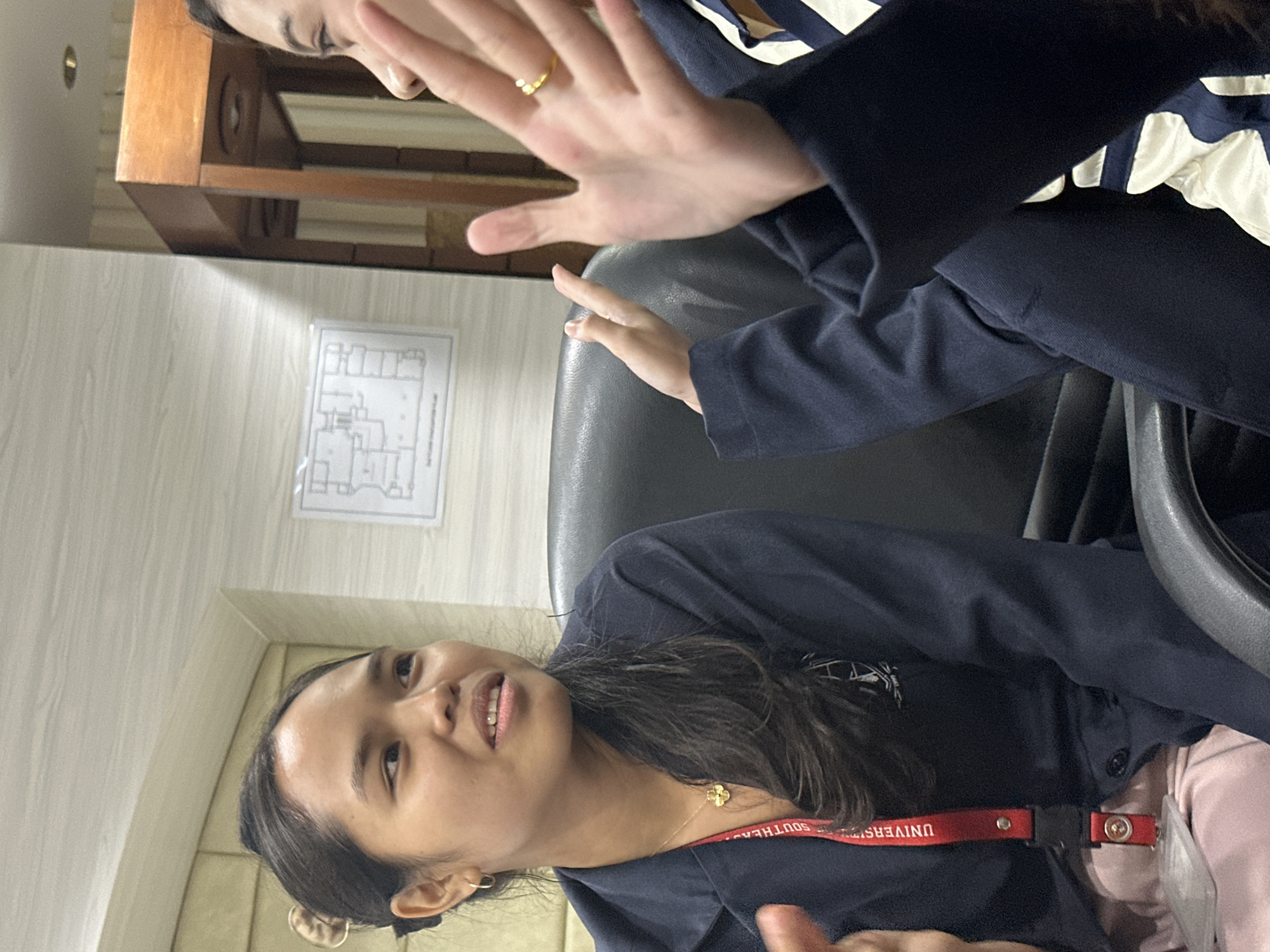 Two young women are engaged in conversation, seated in dark office chairs. The woman on the left, facing the camera, has dark hair pulled back, a gold necklace with a clover pendant, and is wearing a navy blue jacket over a pink garment. A red lanyard with text and a badge is visible around her neck. The woman on the right, whose face is partially visible, is gesturing with her hands and wears a navy jacket with white and blue stripes on her inner garment. She has a gold ring on her right hand. The background includes a light-colored, textured wall with a framed diagram, and a wooden furniture piece. The scene appears to be an indoor office setting within Davao City, Philippines, possibly a university or corporate office, suggested by the lanyard details.