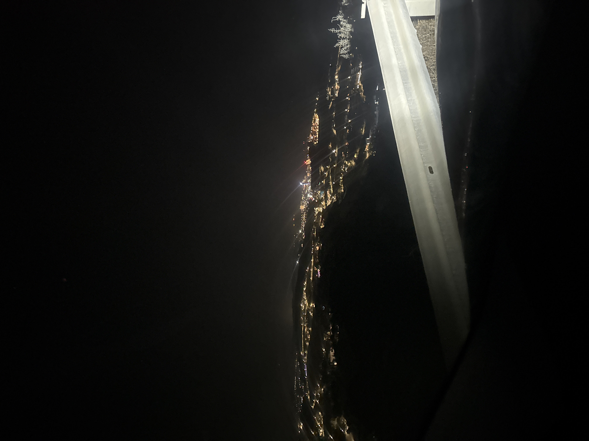 An aerial nighttime view captures the illuminated cityscape of Agios Tychona Community, Cyprus, spread across rolling hills. The darkness of the night sky is punctuated by the warm glow of numerous lights from buildings and streetlights, creating a tapestry of illumination. In the foreground, a reflective metal guardrail, likely from a vantage point such as a road or balcony, runs horizontally across the lower portion of the frame, partially obscuring the view of the city below. The guardrail is textured with a light-colored, possibly gravelly, surface visible behind it. The cityscape itself extends into the distance, with clusters of lights indicating a developed area. The overall scene conveys a sense of urban sprawl against a natural, darkened landscape.