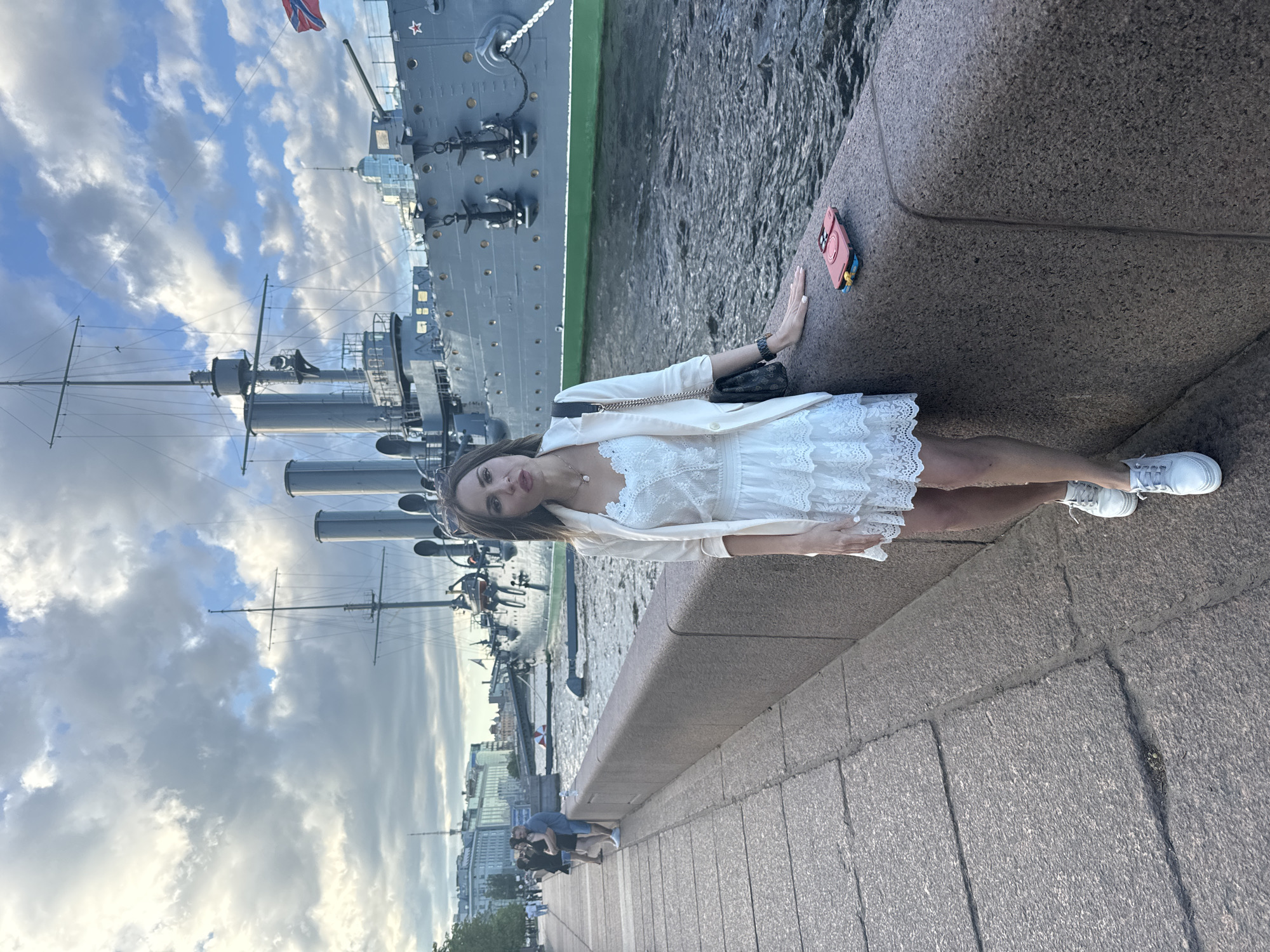 A woman stands on a granite embankment facing the camera. She is wearing a white lace dress, a light-colored blazer, and white sneakers. A black handbag is slung over her shoulder. Next to her on the embankment is a pink phone case. Behind her is a large, grey warship, likely the Aurora cruiser, docked in a body of water. The ship has several smokestacks and a flag at the stern. In the background, buildings and trees line the waterfront, and the sky is partly cloudy. The scene appears to be a quay in Saint Petersburg, Russia, specifically along the Neva River. Several people can be seen walking along the embankment in the distance.
