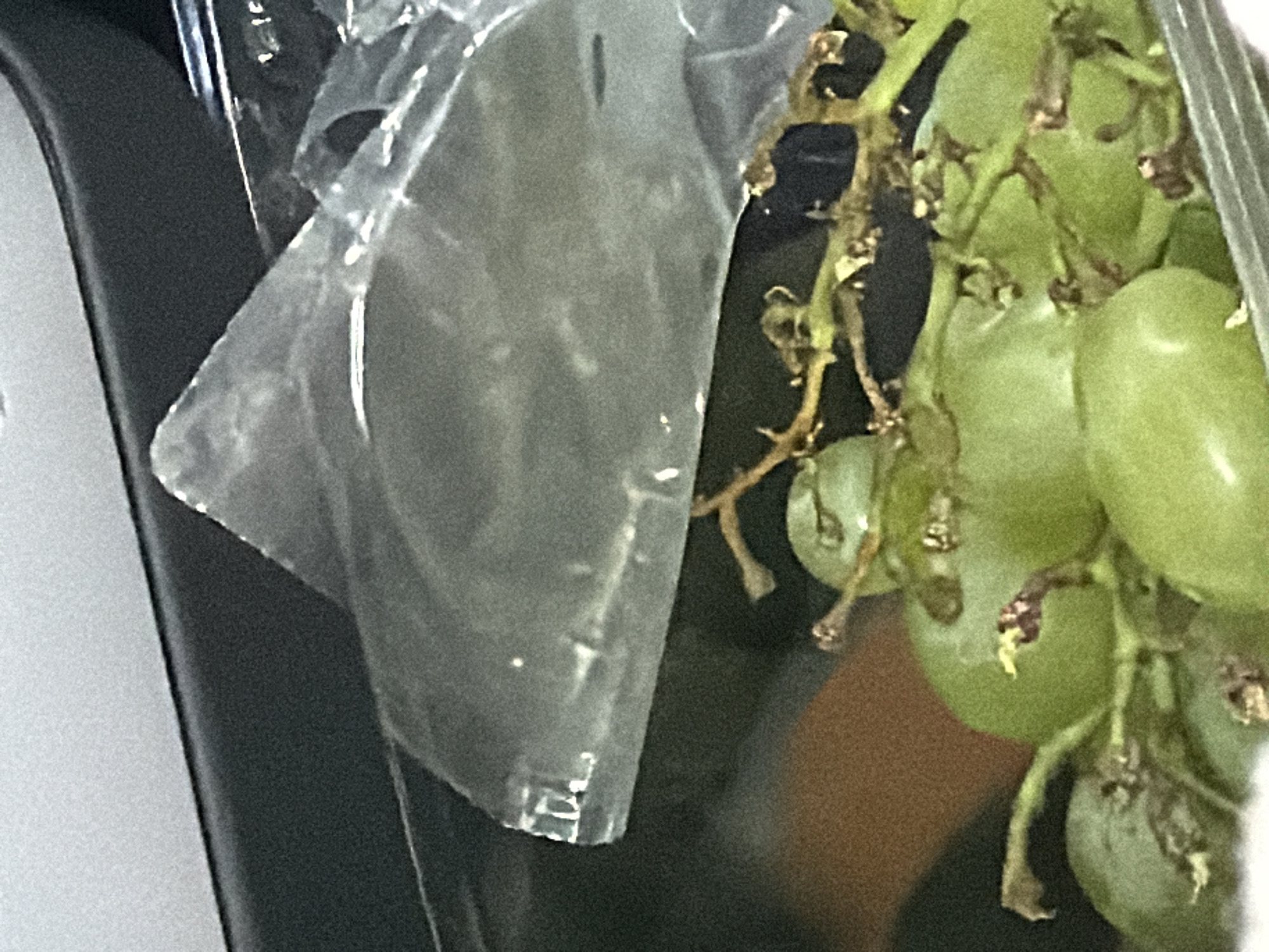 A bunch of green grapes is in a clear plastic container. The container is on a dark surface. The grapes have brown stems and some have small brown spots on their skin. The background is blurry, but it appears to be indoors. There is a grey surface above the container, and a brown surface below it. The image does not provide any information about the location of Löftaskog, Sweden.