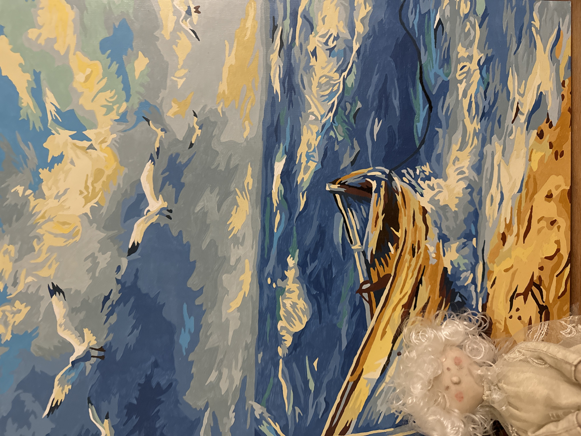 The image shows a close-up of a painting depicting a seascape with flying birds on the left and a boat near the right. The painting is rendered in a flat, illustrative style with bold blocks of color. A porcelain doll with white curly hair rests against the painting's lower right corner.