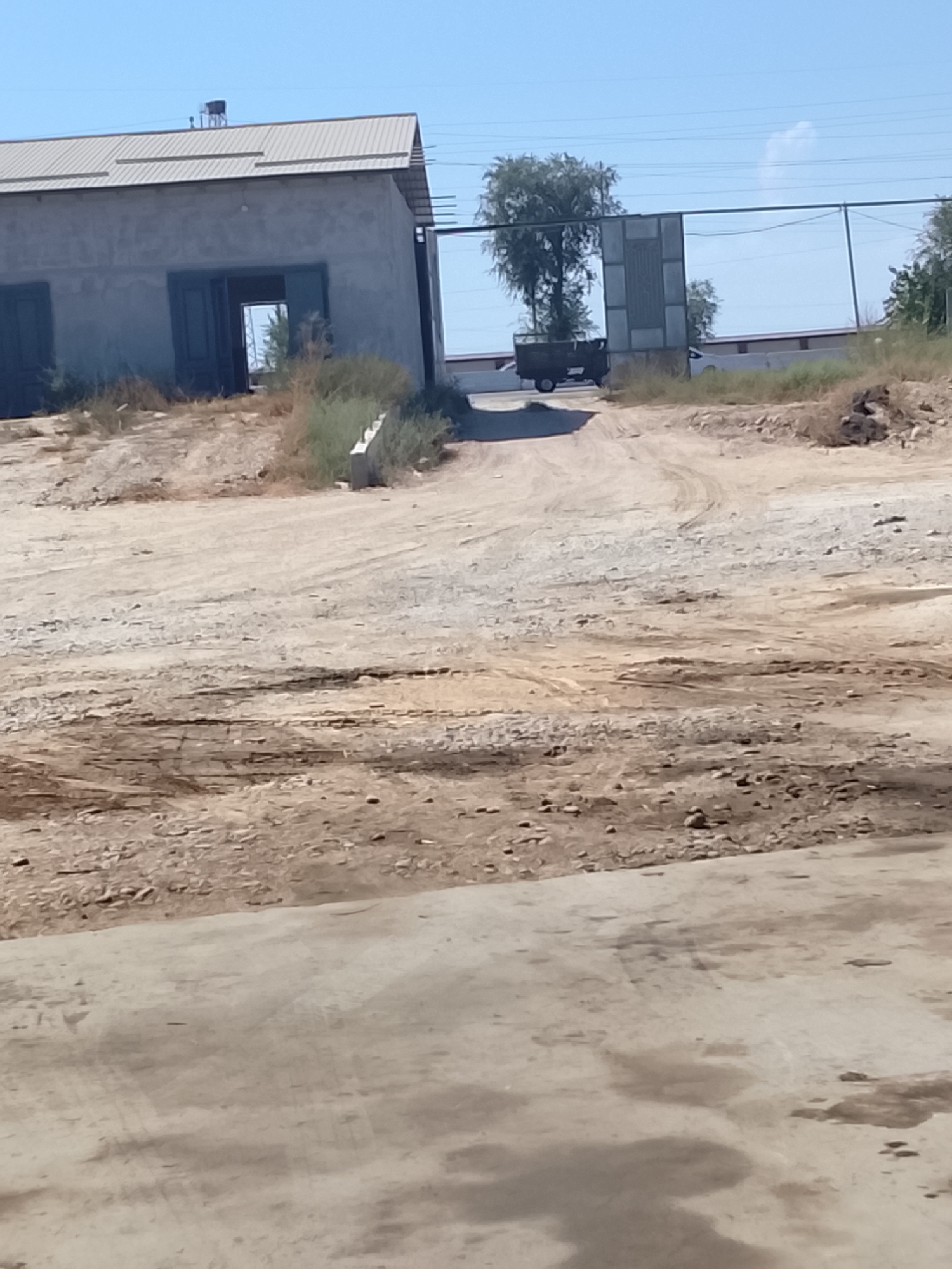 The image shows an outdoor scene with a building and a dirt road. The building appears to be a warehouse or industrial facility with a grey, corrugated metal roof and light-colored walls. There are large open doorways on the front of the building. In front of the building, a dirt road leads up to an open gate or entrance. A small, dark-colored truck is visible on the road, partially obscured by a gate structure. The area is dry and dusty, with sparse vegetation. The sky is clear and blue. The scene is consistent with an industrial or agricultural area in Sabir Rakhimov, Uzbekistan.