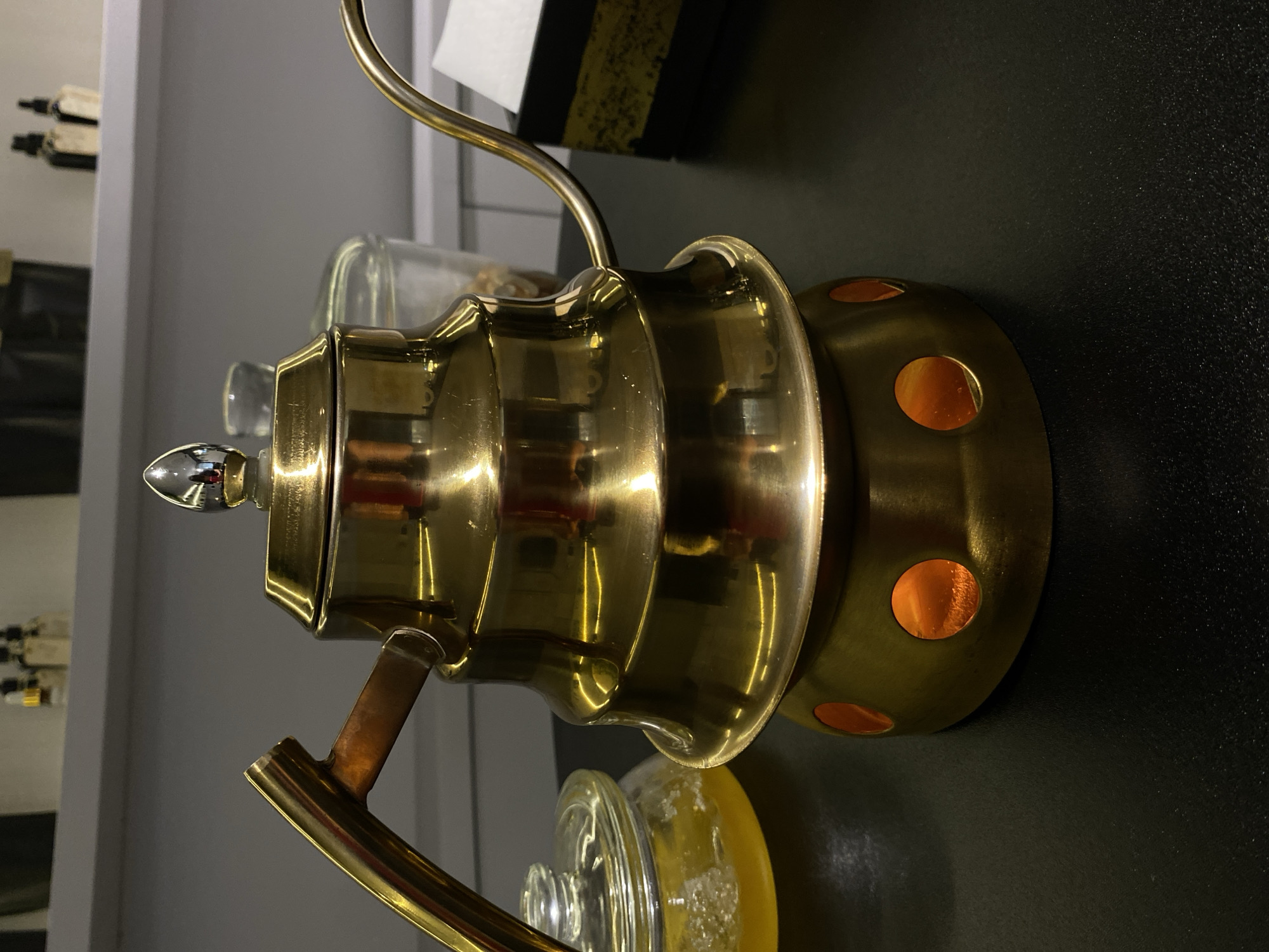 A close-up, eye-level shot focuses on a gleaming, brass-colored gooseneck kettle situated on a black surface. The kettle has a distinctive tiered shape and a copper-colored handle. It rests on a circular brass base with circular cutouts that reveal an orange glow, suggesting a heat source within. To the left of the kettle, a glass jar with a lid contains a yellow liquid with small particles. In the background, out of focus, a shelf holds several small glass bottles with dark caps. The environment appears to be an indoor setting, possibly a cafe or kitchen, with muted lighting that highlights the reflective surfaces of the kettle. The image does not contain any elements to suggest a location in Votkinsk, Russia.