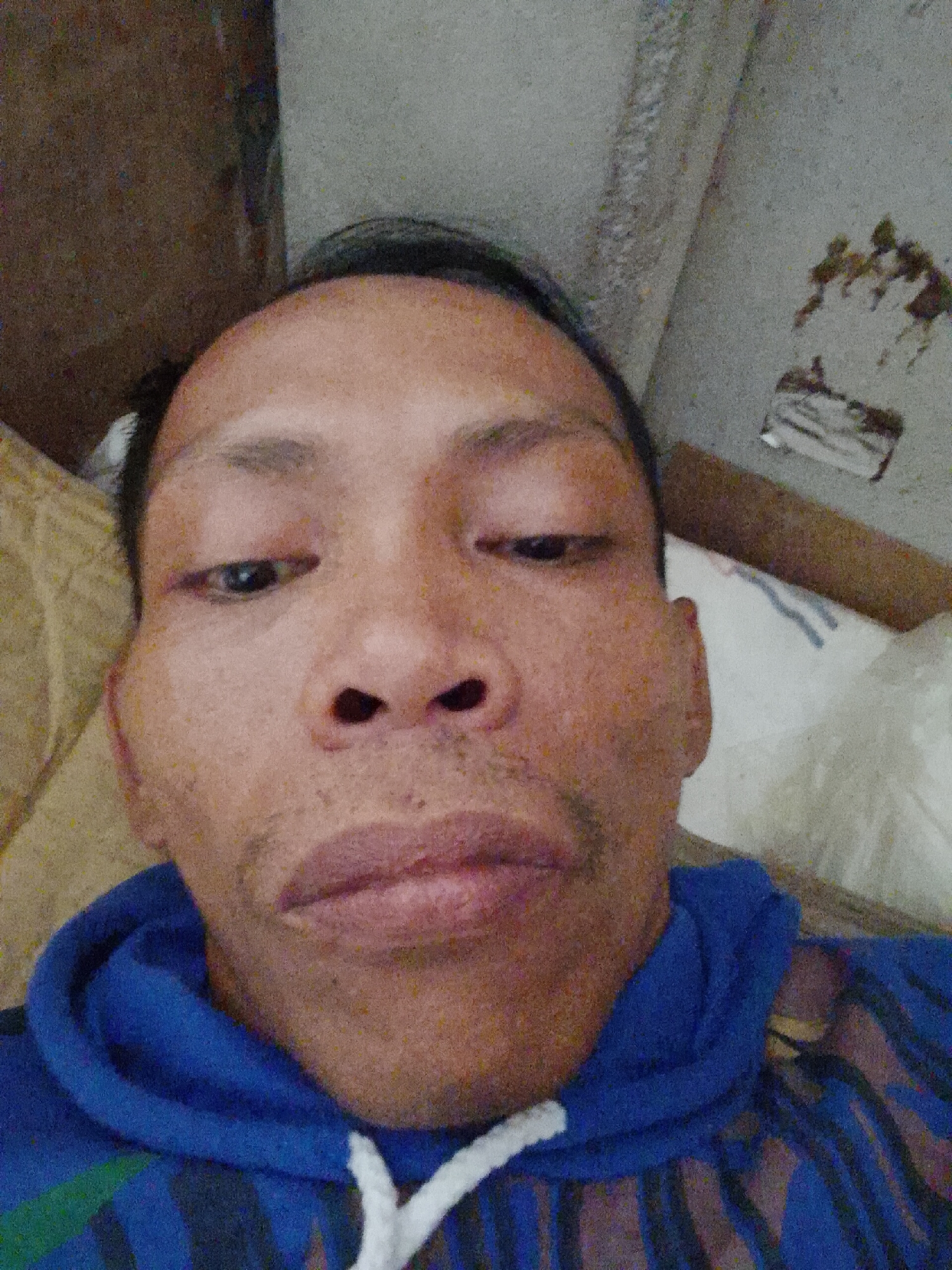 A close-up, selfie-style image depicts a person with dark hair and complexion, wearing a blue and striped hoodie. The person's face is the primary subject, showing details of their eyes, nose, and mouth. The background is blurry and indistinct but suggests a room interior with a wall and bedding visible. There is no information in the image that indicates the location is in Bukit Ketok, Indonesia. The context lacks specifics to confirm the location or any activities.