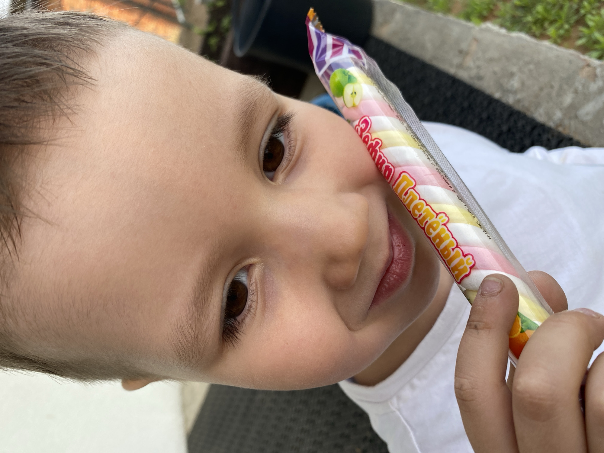 A child's face, with brown eyes and short brown hair, is close to the camera. The child is holding a packaged confection labeled "Zova Pletenyi." The confection appears to be twisted marshmallows. The packaging has images of an apple and citrus fruits, and text is written in Russian. The background shows partial views of a white shirt, a concrete surface, and greenery. The packaging indicates the product location is in the city of St. Petersburg, Russia.
