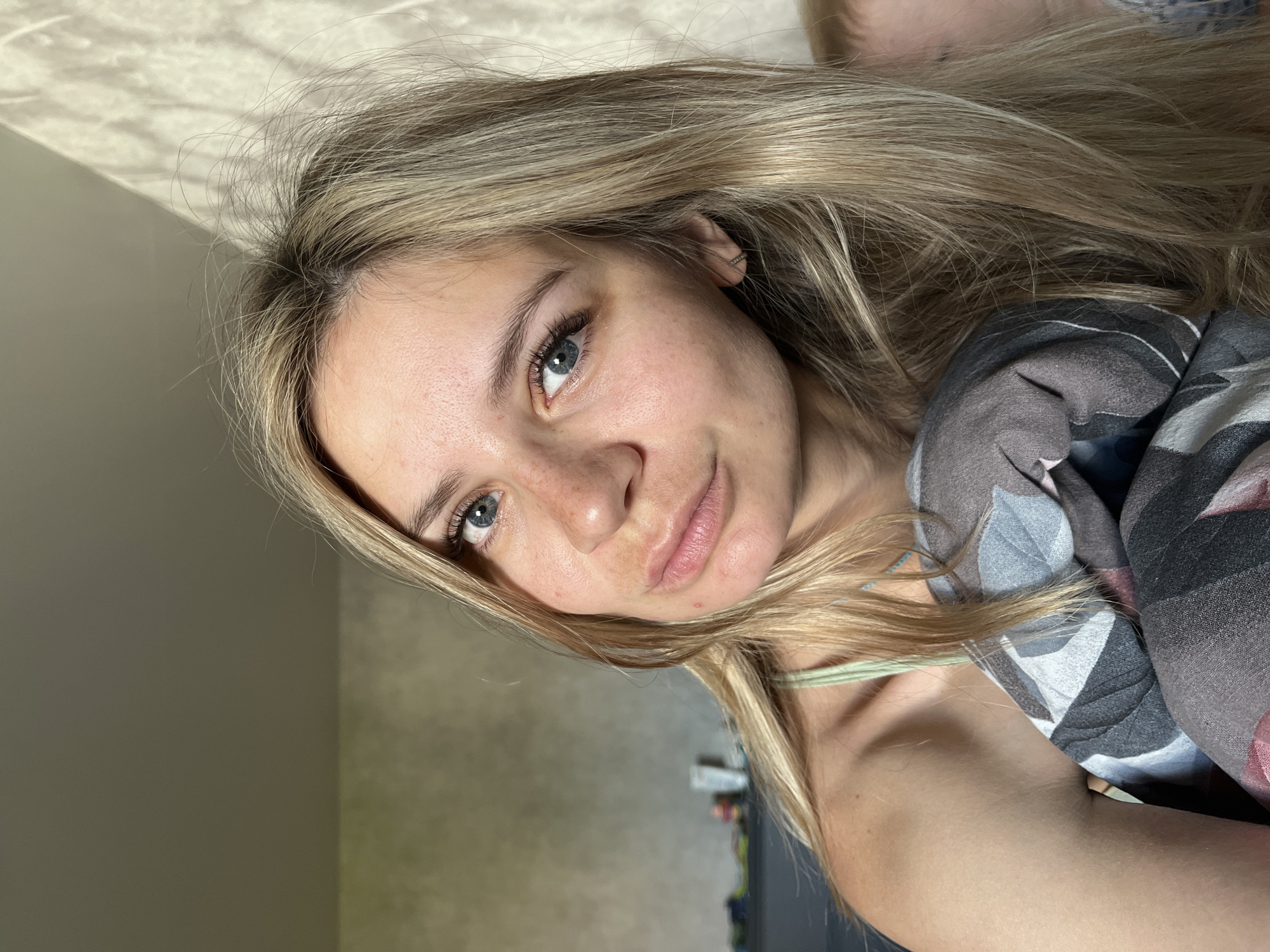 The image depicts a close-up, indoor portrait of a young Caucasian woman with blonde hair and blue eyes, who is looking towards the camera. She has freckles on her nose and cheeks. The woman is wearing a light green camisole and is partially covered by a blanket with a floral pattern. A blurred background suggests an interior setting, possibly a bedroom, with a wall and some furniture visible. The lighting indicates natural light coming from the right side of the frame. There is no discernible connection to the city of Chaikovsky, Russia, or any specific urban environment.