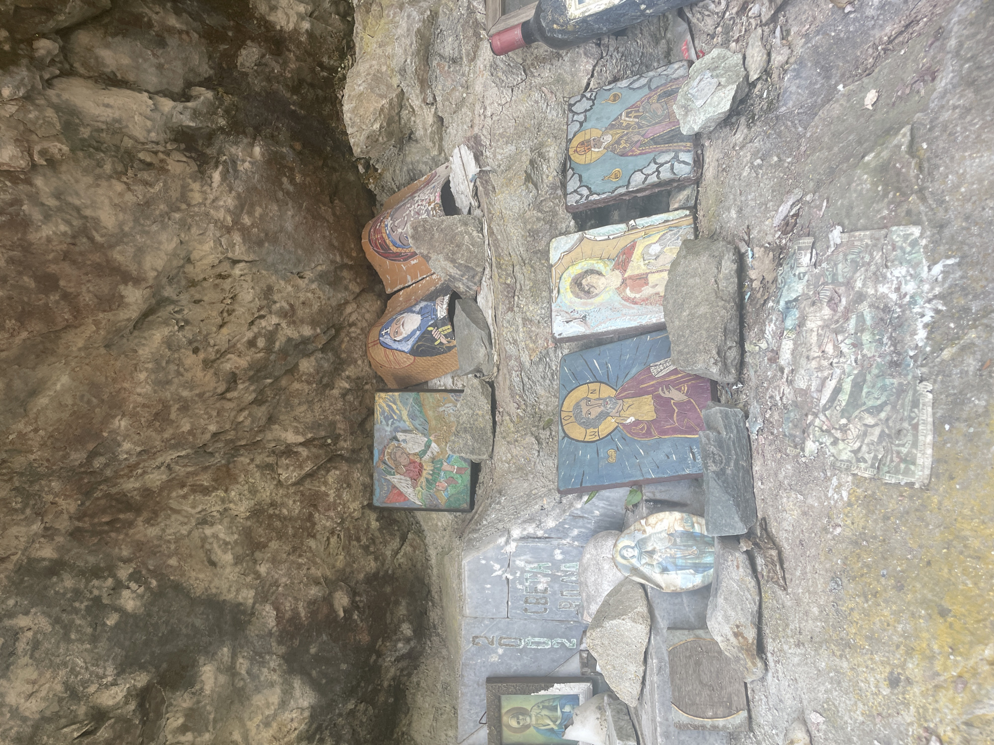 The image depicts a rock face or grotto adorned with religious icons, pictures, and other items. The location appears to be a natural niche or small cave within a rock formation, possibly outdoors. The icons and pictures are a mix of traditional Orthodox Christian imagery. The objects are arranged in an informal, almost haphazard manner, suggesting that the place may be a spontaneous shrine or a place of personal devotion. The setting seems to be in the city of Sofia, Bulgaria.