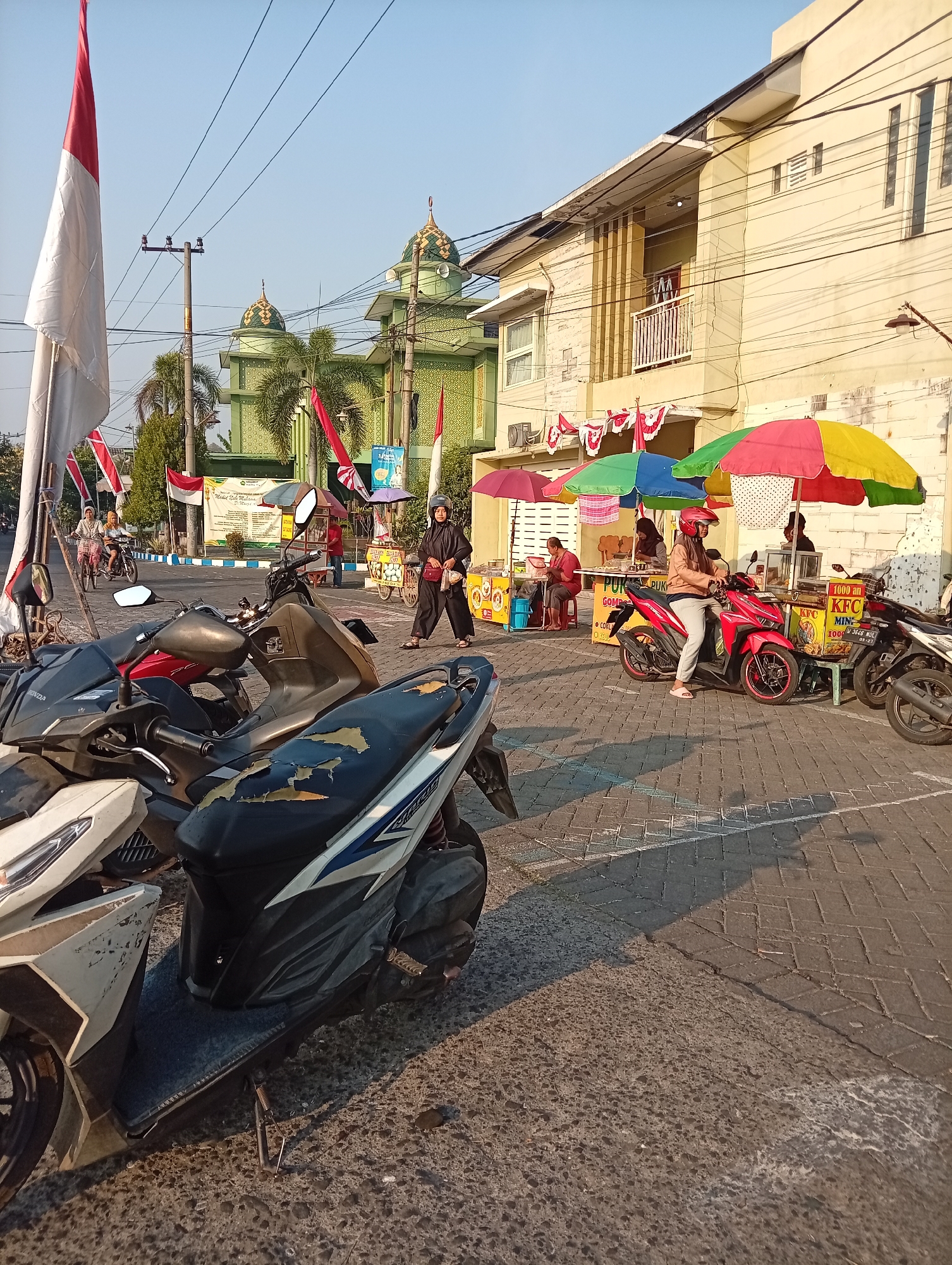 In Bligo, Indonesia, the scene captures an outdoor marketplace with a street and a building in the background. The street is paved with interlocking bricks, and several motorcycles are parked in the foreground. A large flag, red and white, is prominently displayed on the left side of the image. Behind the motorcycles, a row of vendors operates under colorful umbrellas. One vendor is selling food from a cart, while others are attending to customers. A woman in a black hijab and black clothing is seen walking away from the food cart, carrying a bag. Further in the background, a green building with domes, likely a mosque, is visible, adorned with Indonesian flags. The sky is clear and blue, indicating a sunny day. The overall atmosphere suggests a bustling local market with a blend of commerce and cultural elements.
