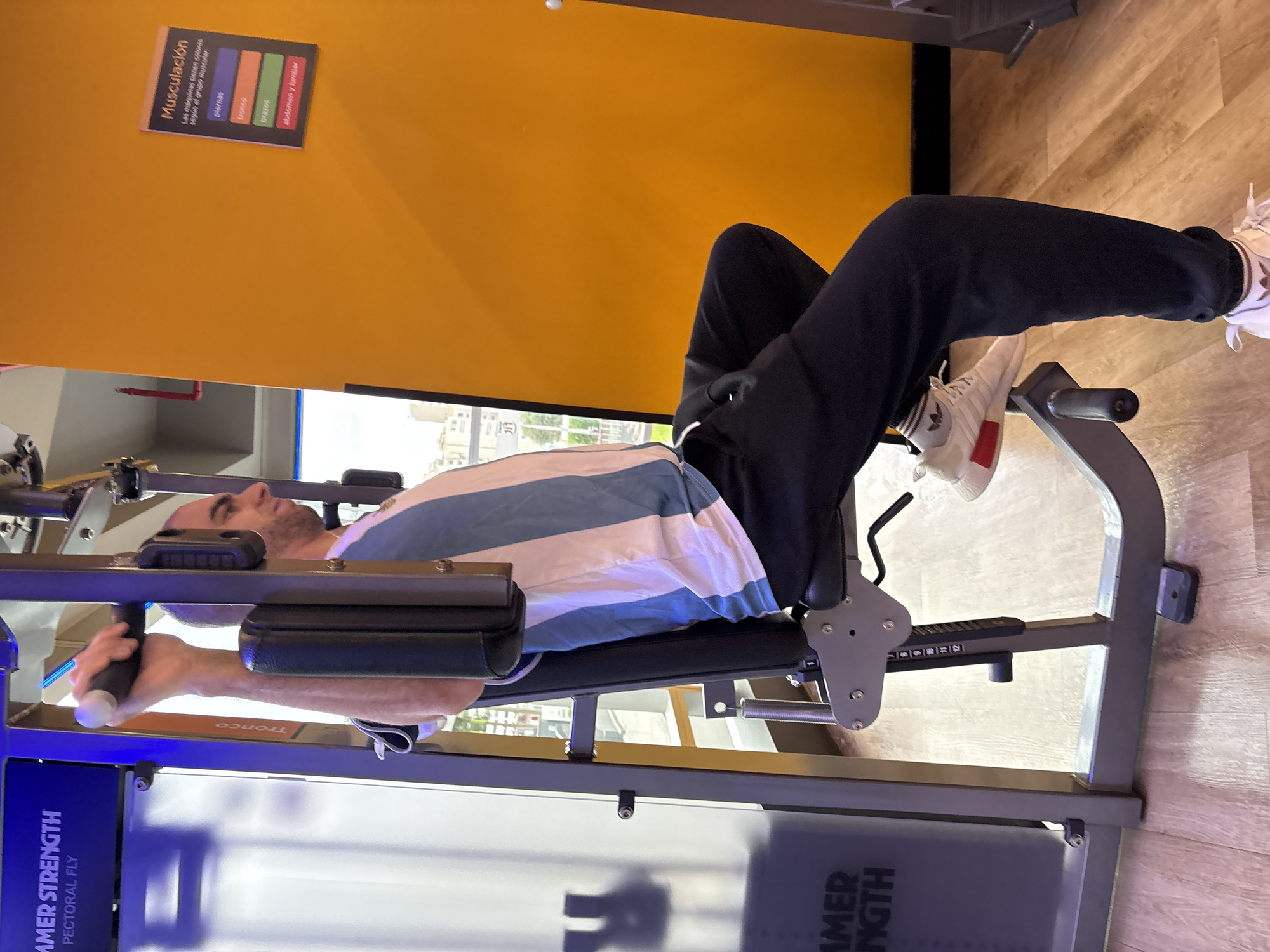 In a gym setting in San Isidro, Peru, a man with a shaved head is seated on a weight machine, performing a chest exercise. He is wearing a white t-shirt with blue vertical stripes and black sweatpants. His right hand grips a handle of the machine. The machine is gray and has a blue label that reads "HAMMER STRENGTH PECTORAL FLY." To the right of the man, on an orange wall, is a small sign with the title "Musculación" and color-coded muscle groups, including "piernas" (legs), "tronco" (torso), "brazos" (arms), and "abdomen y lumbar" (abdomen and lower back). The floor is light-colored wood.