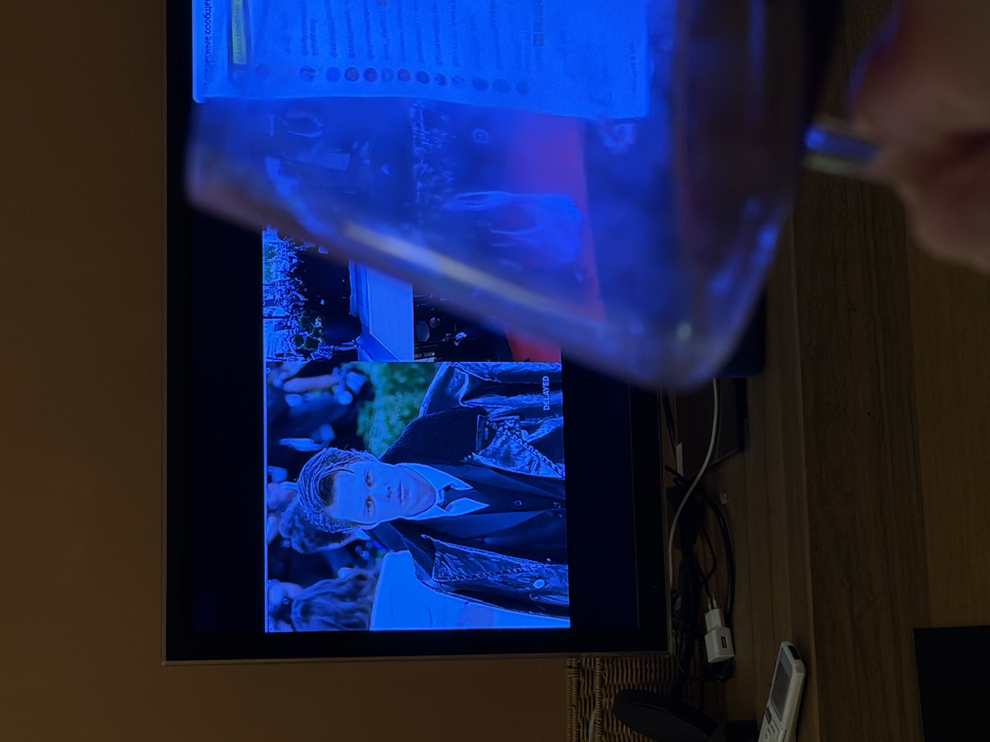 The image shows a television screen displaying a scene with a man wearing a black jacket and a white shirt and tie. The screen is partially obscured by a translucent, possibly plastic, object held up in front of it. Cables and electronic devices are visible near the bottom right of the television. The environment appears to be an interior setting, possibly a home, with a wood-paneled wall and a wooden table surface. There is no indication that this photo was taken in Tel-Aviv, Israel.